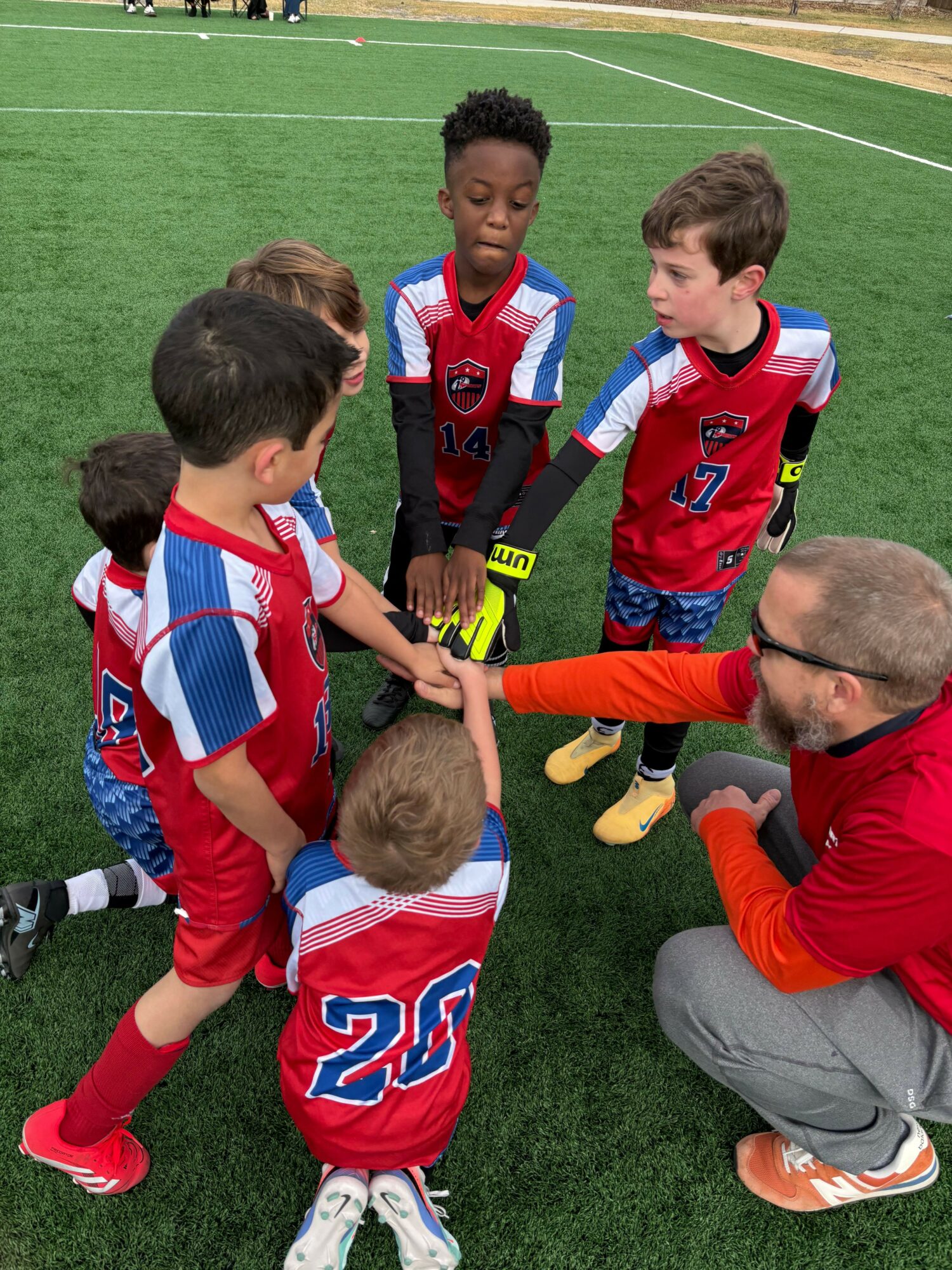 Two young soccer players in red and blue uniforms are on the ground, with a man assisting them on a green field.