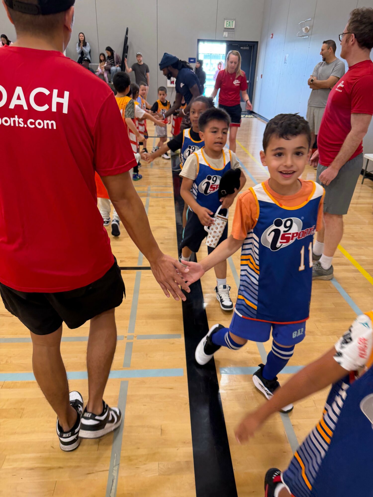 Children in sports uniforms receive instructions from a coach in a gymnasium, with others watching in the background.