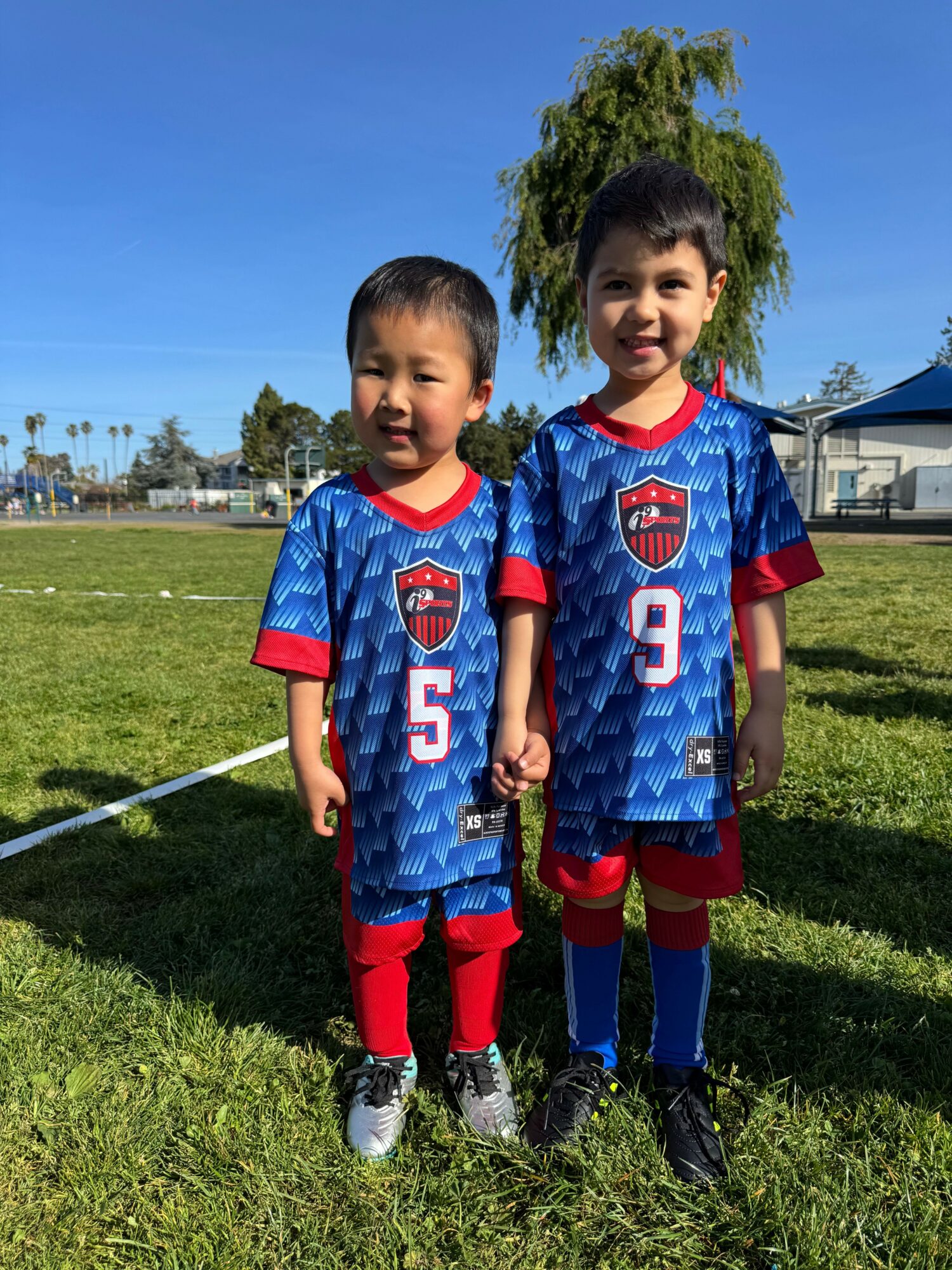 Two children in soccer uniforms standing on grass field, smiling, with trees and blue sky in background.