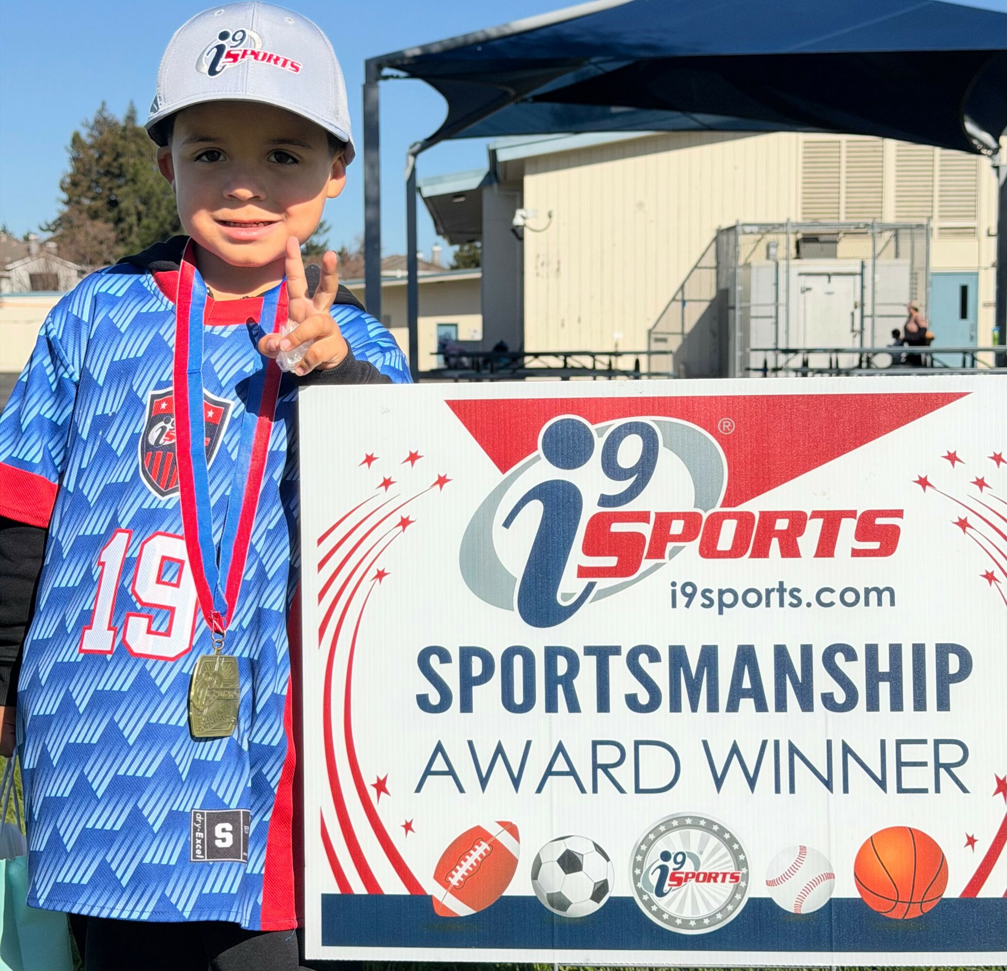 Young boy in sports jersey and cap holding a medal, standing next to a sportsmanship award sign outdoors.