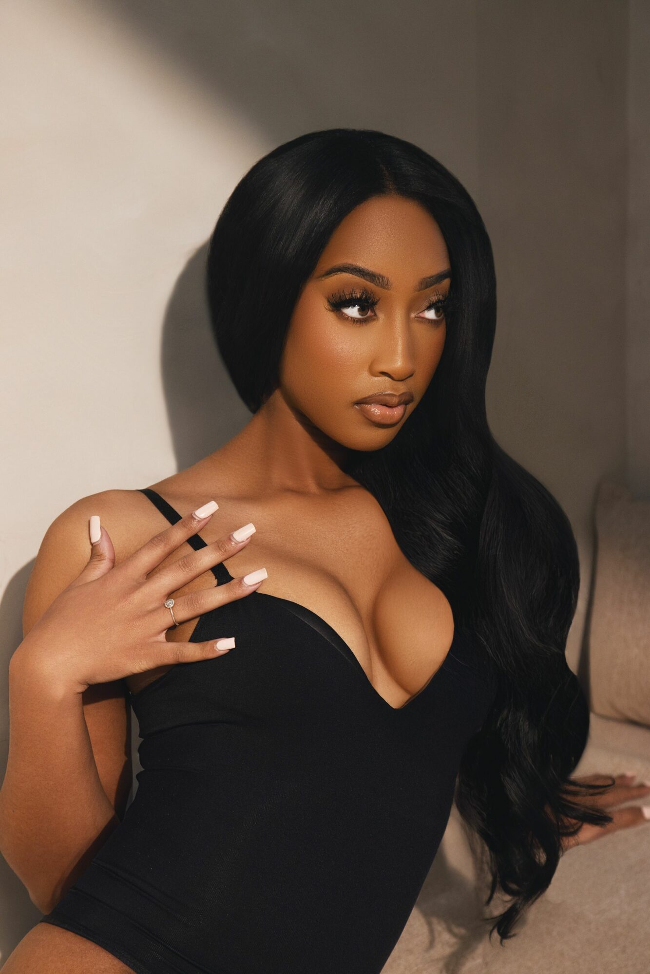 Woman with long black hair, wearing a black strapless top, looking to the side, with hand near face, neutral background