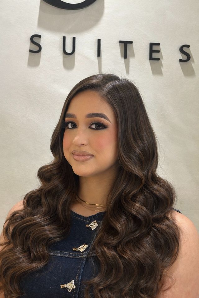 Young woman with long, wavy hair and makeup, standing in front of a wall with the word 'SUITES' displayed.