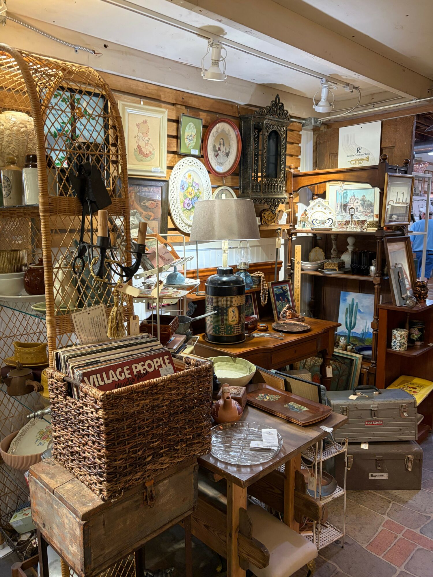Room filled with various antique and vintage items, including furniture, lamps, and decorative objects, arranged on tables and shelves.