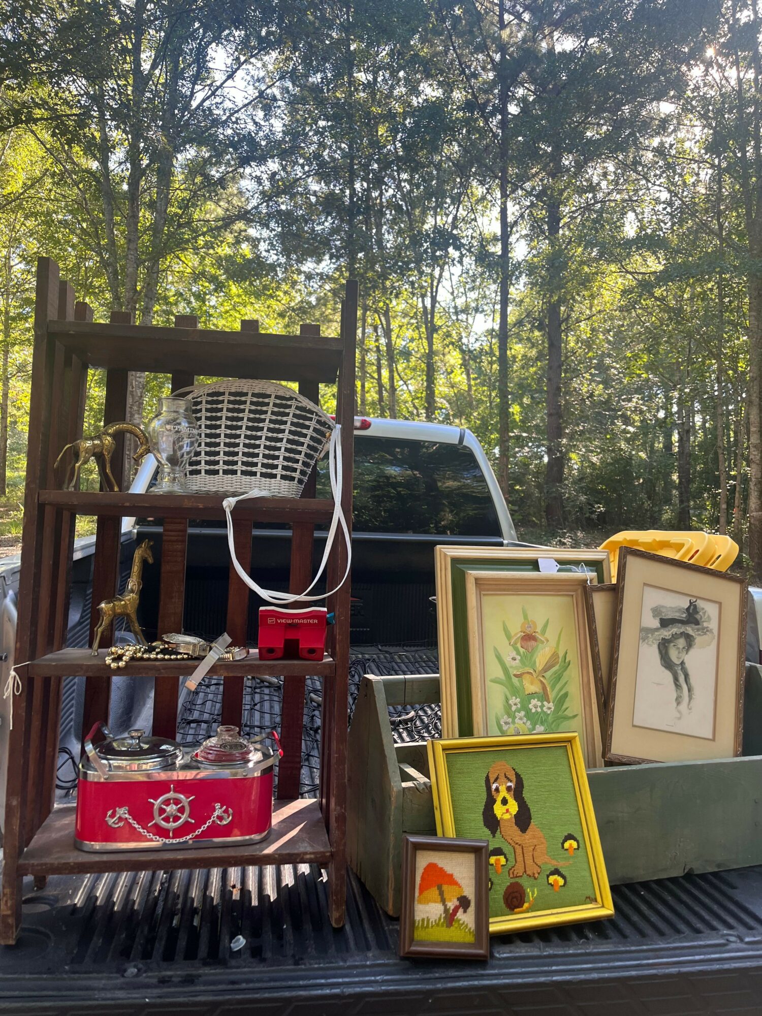 Outdoor scene with framed artwork and decorative items on a table, trees in the background, and a chair nearby.