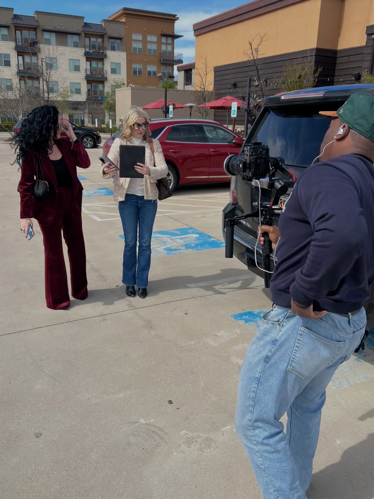 Two women being filmed by a man with a camera in a parking lot, with buildings and cars in the background.