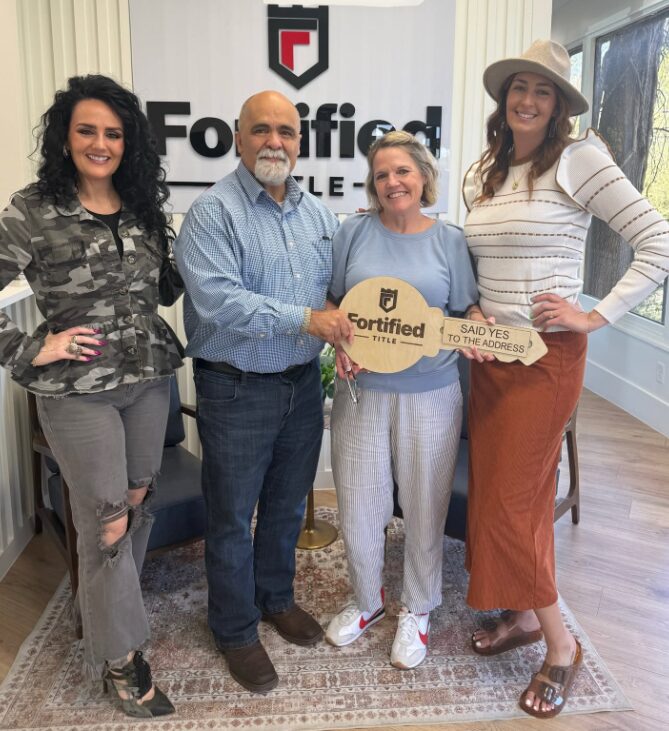 Four people standing indoors, holding a large key and a sign, smiling, with a logo and text in the background.