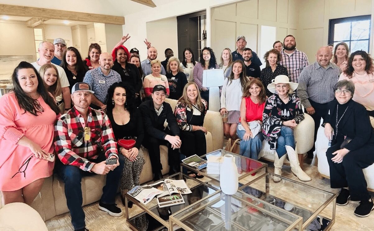 Group of people gathered indoors, some sitting and some standing, smiling for a photo, with tables and windows in the background.