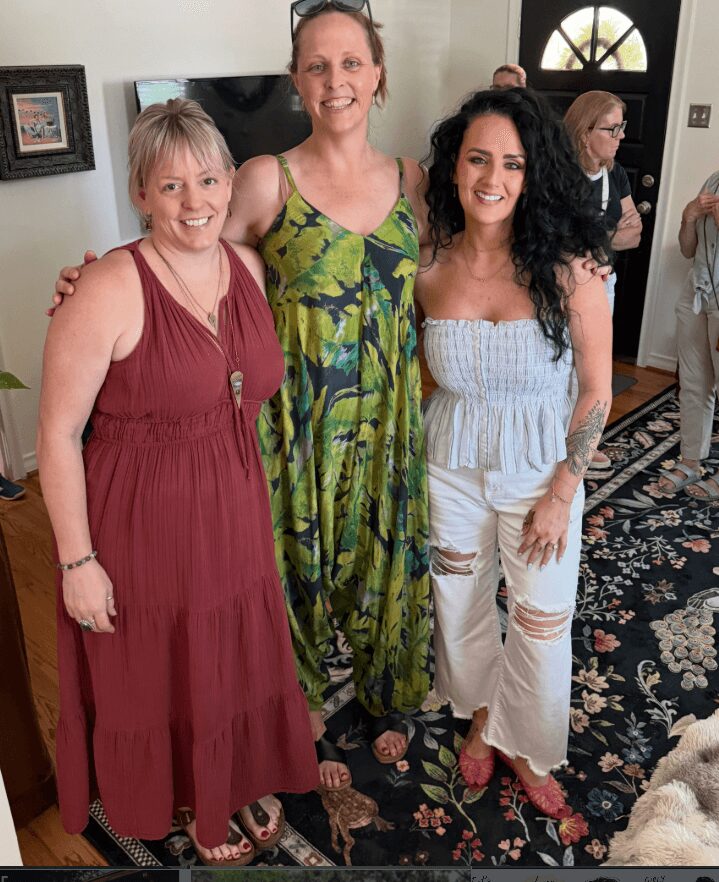 Three women standing indoors, smiling, with a patterned rug and a door in the background.
