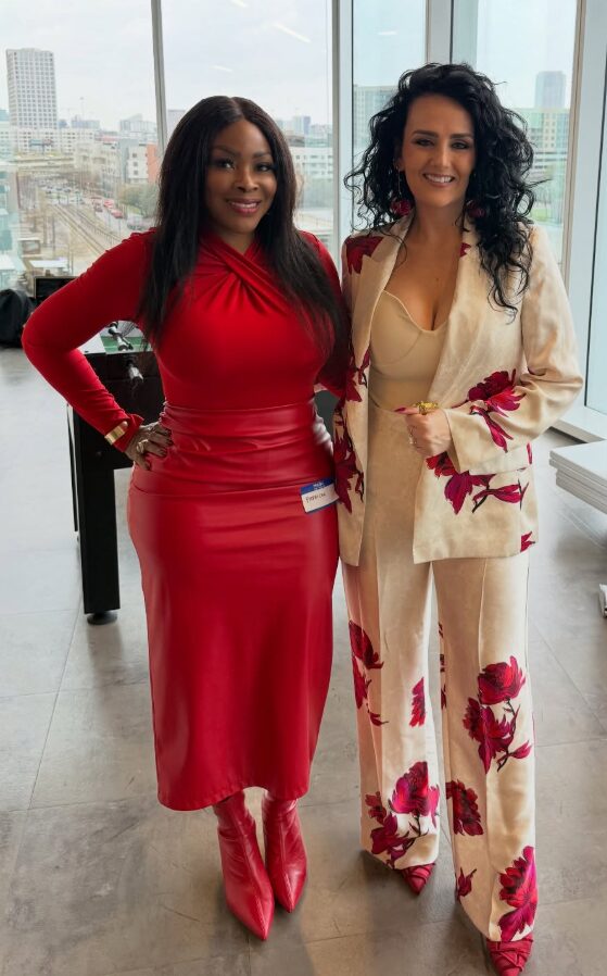 Two women standing indoors with city view, smiling, one in a red dress and boots, the other in a floral suit.