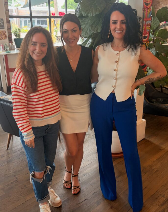 Three women standing indoors, smiling, with plants and windows in the background.