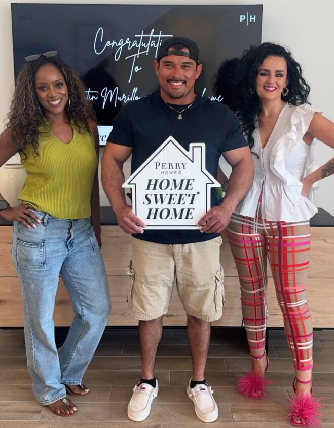 Three people standing indoors, smiling, with a sign that says 'Home Sweet Home,' in front of a blackboard with writing.