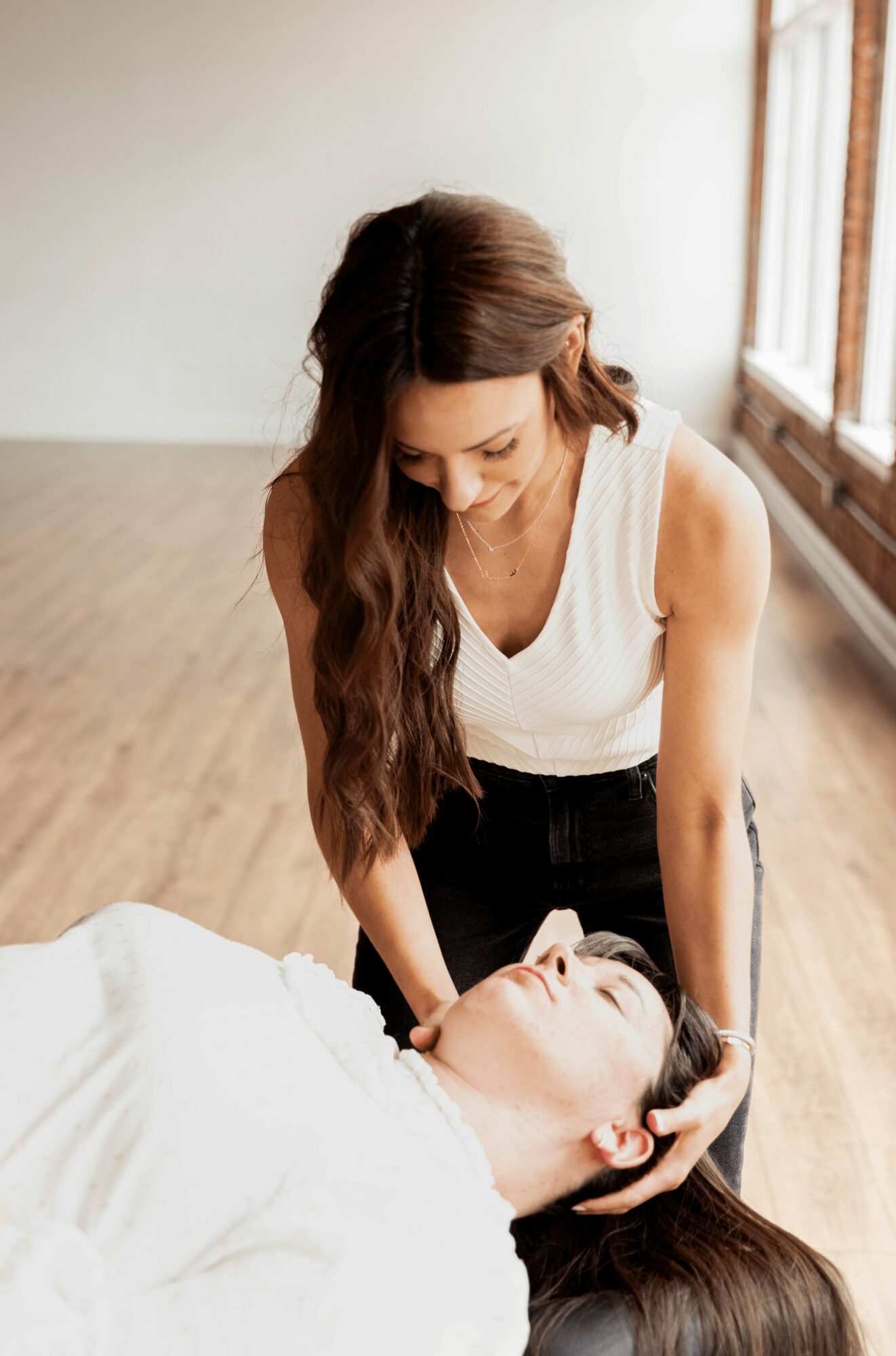 Two women in a room with wooden floors and large windows, one standing and the other lying down, engaging in a caring gesture.
