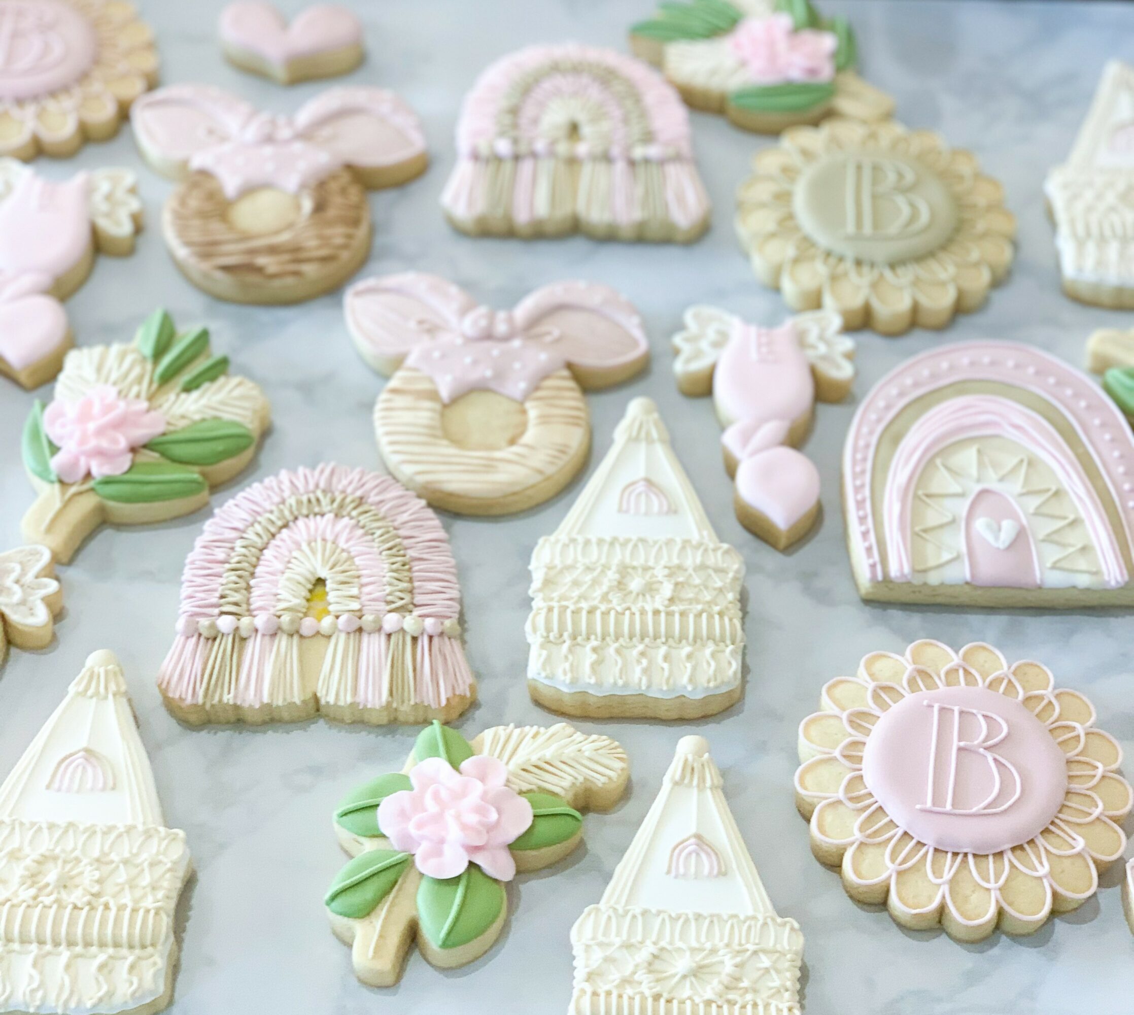 Assorted pastel-colored cookies with bows, flowers, rainbows, and monogrammed designs on a light background.