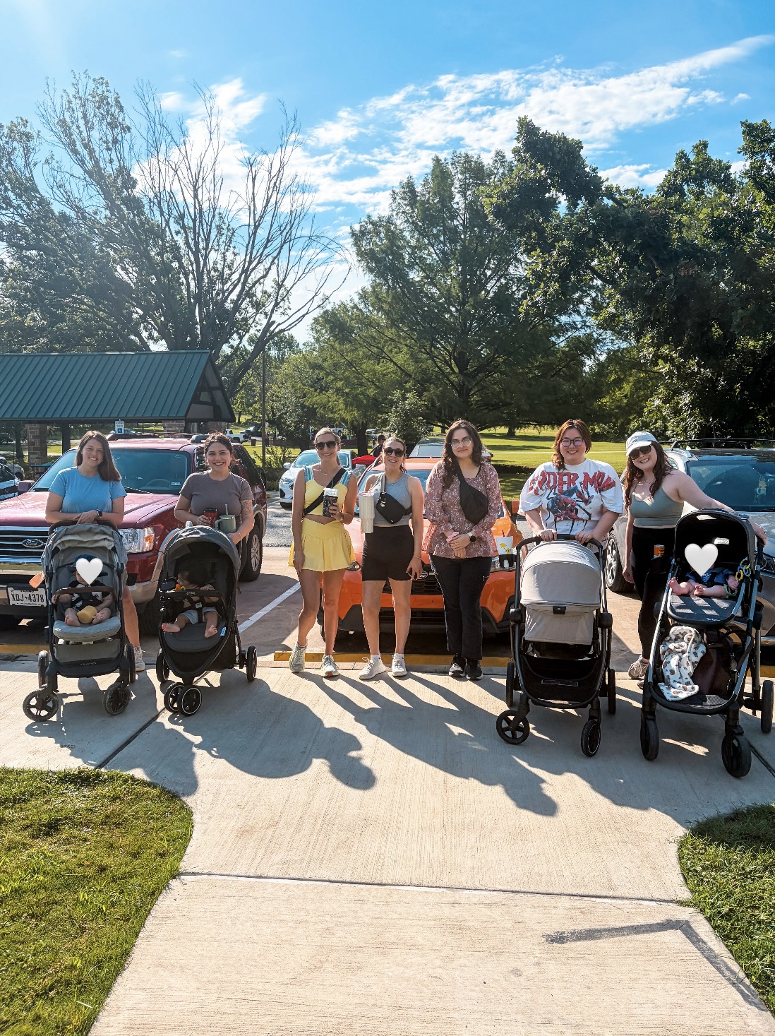Group of seven women standing outdoors with strollers, trees, and parked cars in the background, sunny day with blue sky.