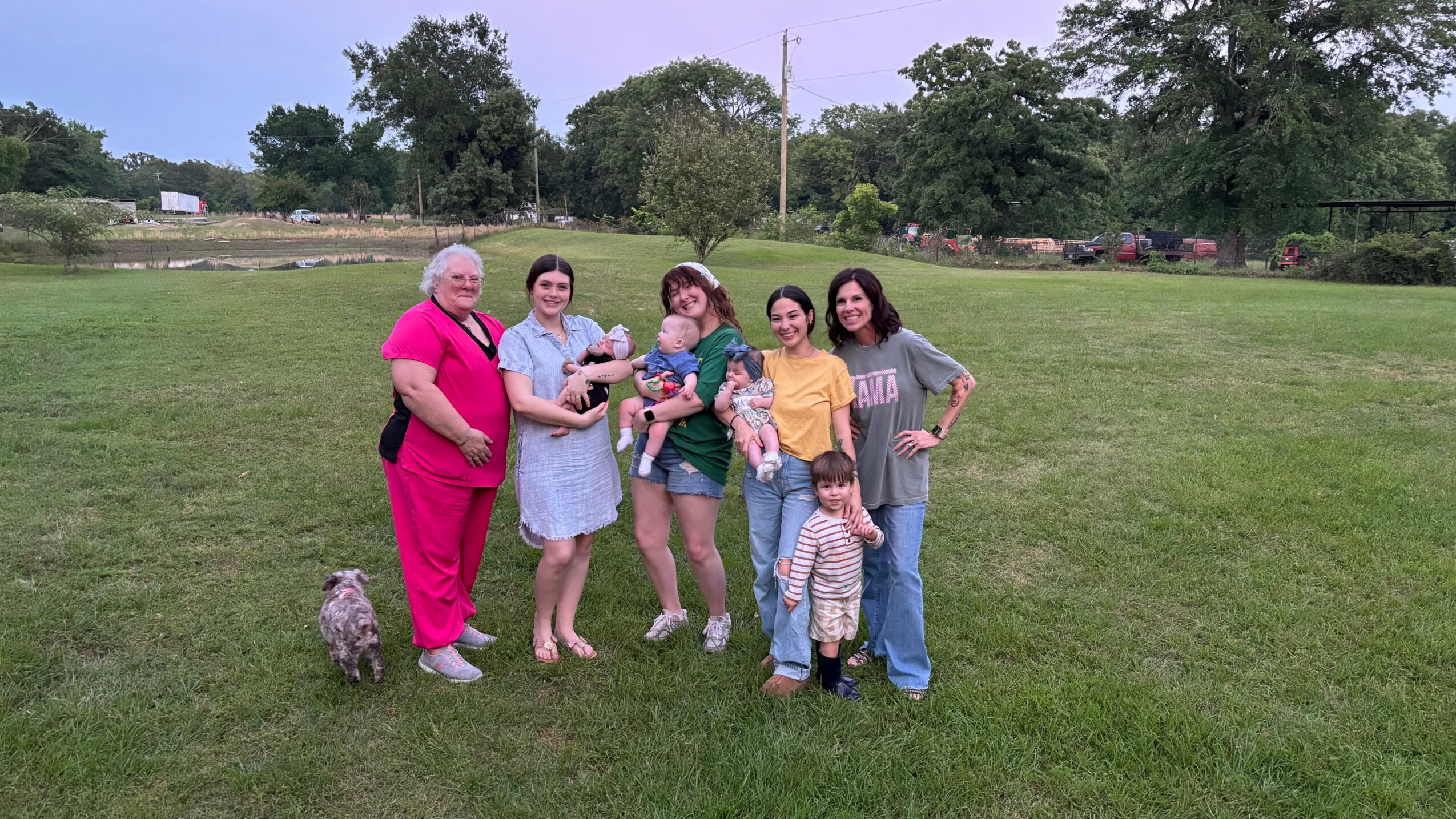 Group of seven people standing on grass in a park, with trees and a fence in the background.