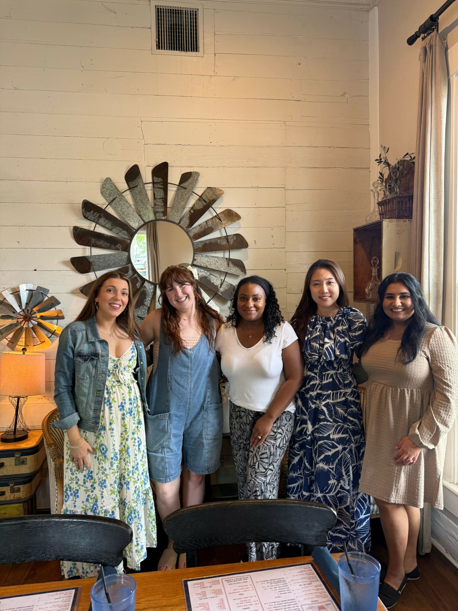 Five women stand together in a room with a decorative mirror on the wall, a table with papers, and a colorful umbrella decoration.