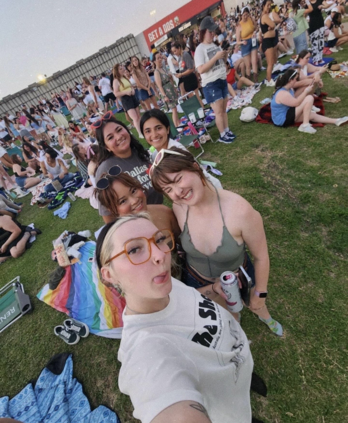 Group of five young women taking a selfie on grass at outdoor event with many people in background.