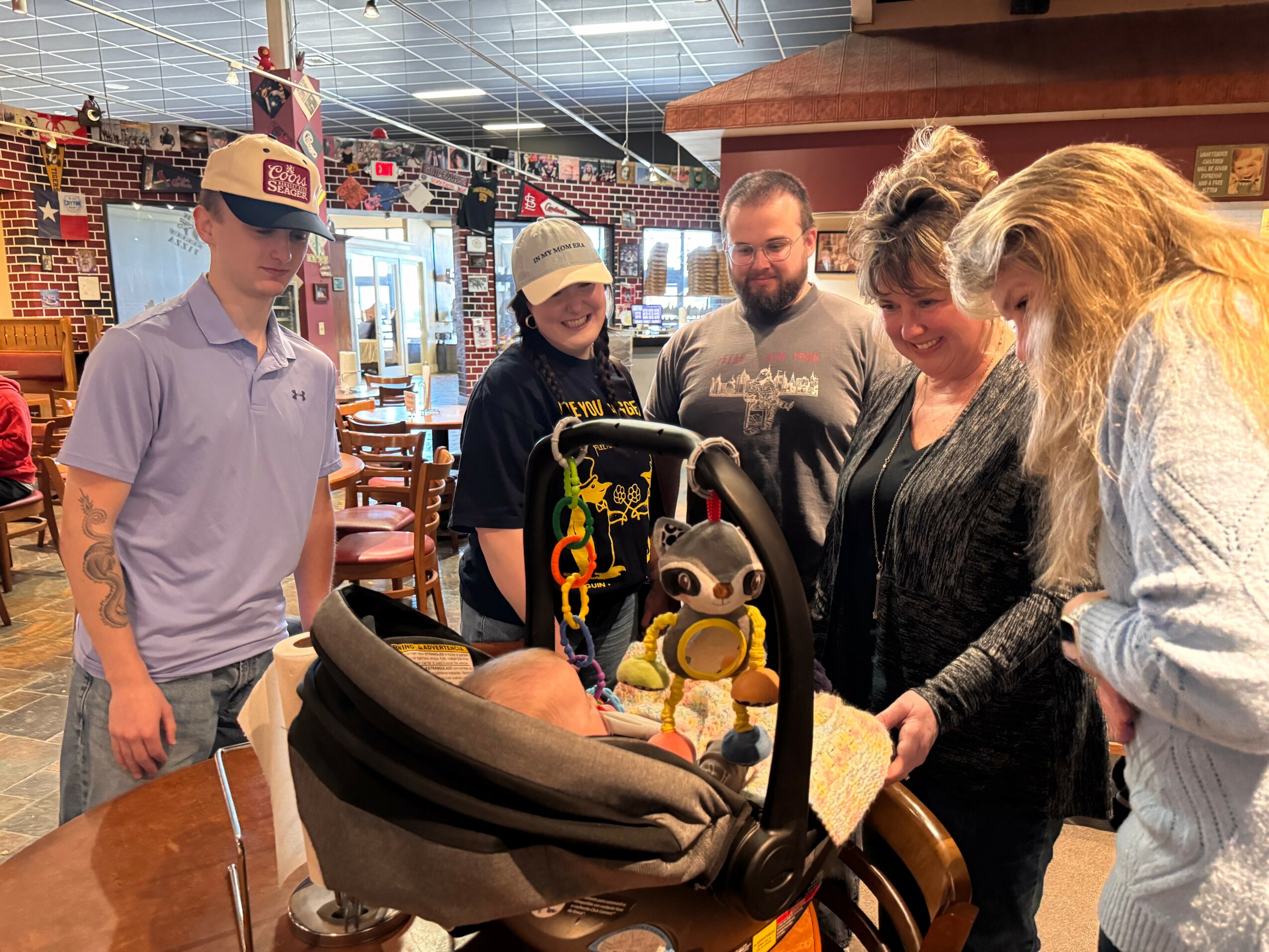 Group of five people gathered around a baby in a car seat inside a store, smiling and talking.