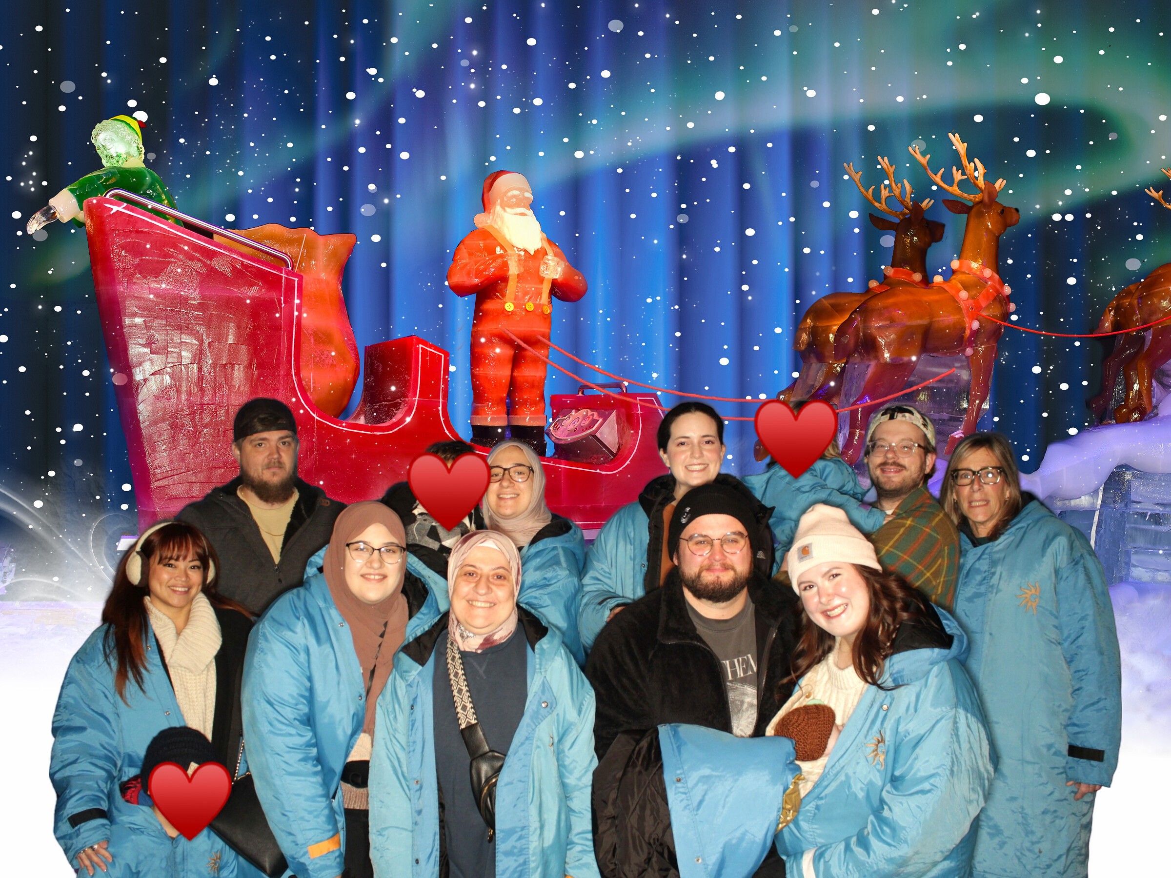 Group of people in blue jackets posing in front of a festive holiday display with Santa, reindeer, and northern lights.
