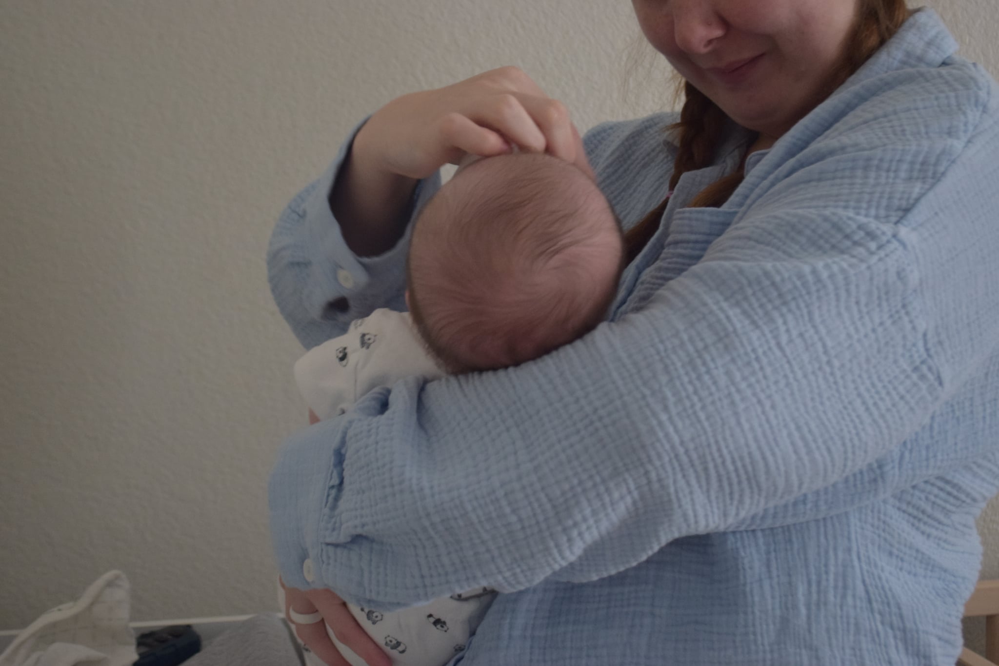 Person holding a baby, gently touching the baby's head, wearing a light blue garment, against a plain background.