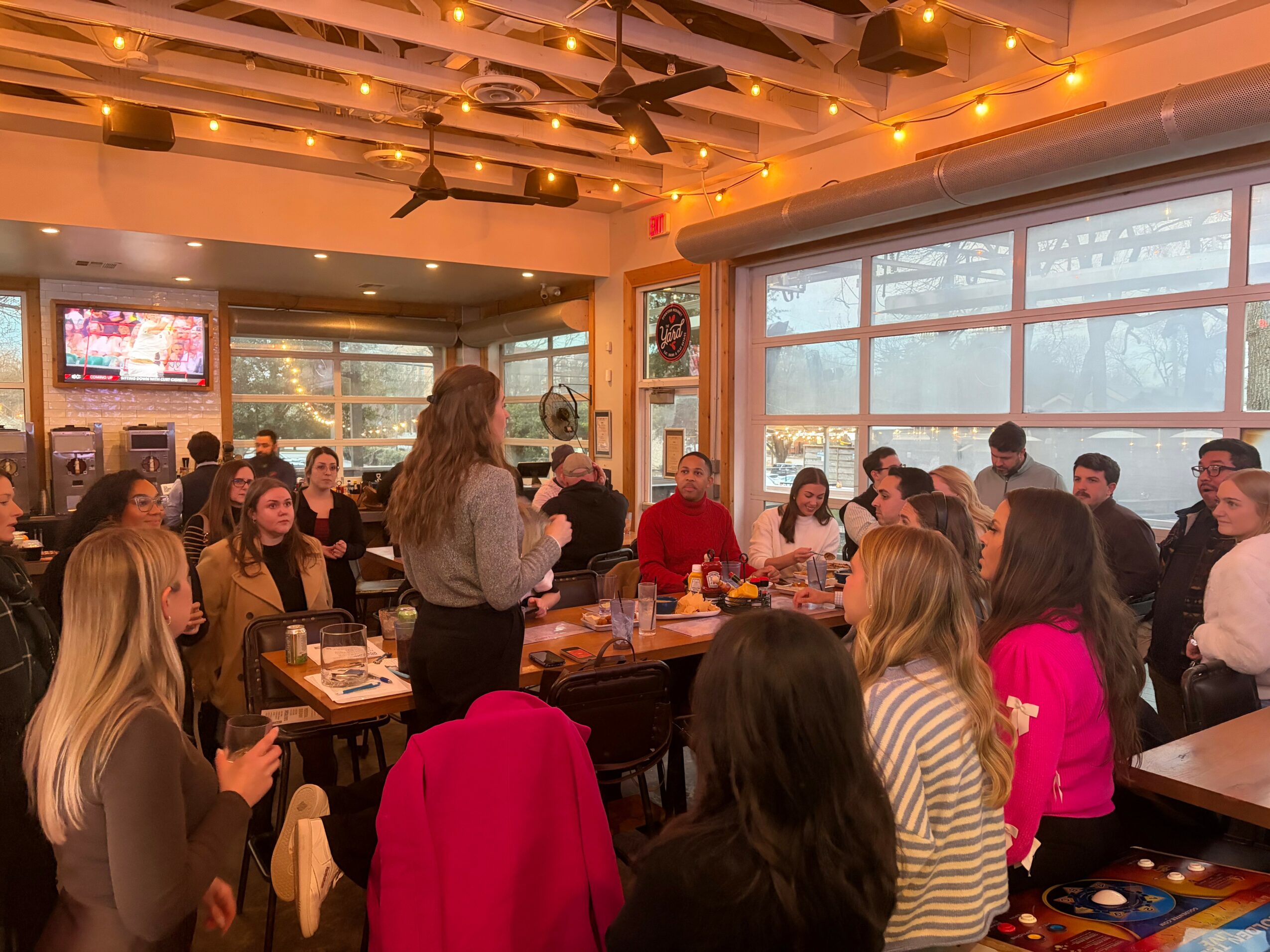 Group of people gathered around a table in a restaurant, listening to a woman speaking, with large windows and warm lighting.