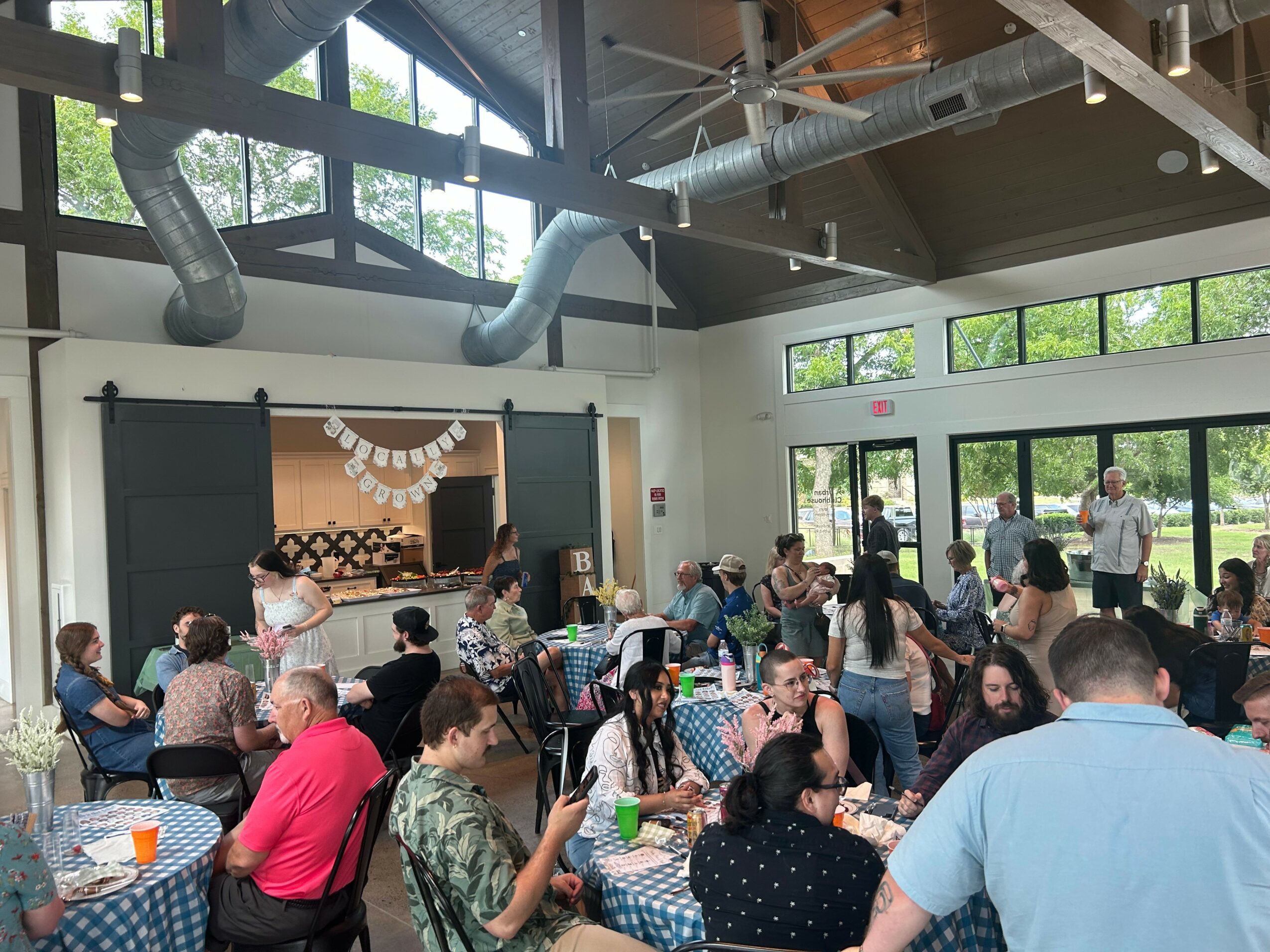 Indoor event with people seated at tables, some standing, in a bright room with large windows and a stage at the front.