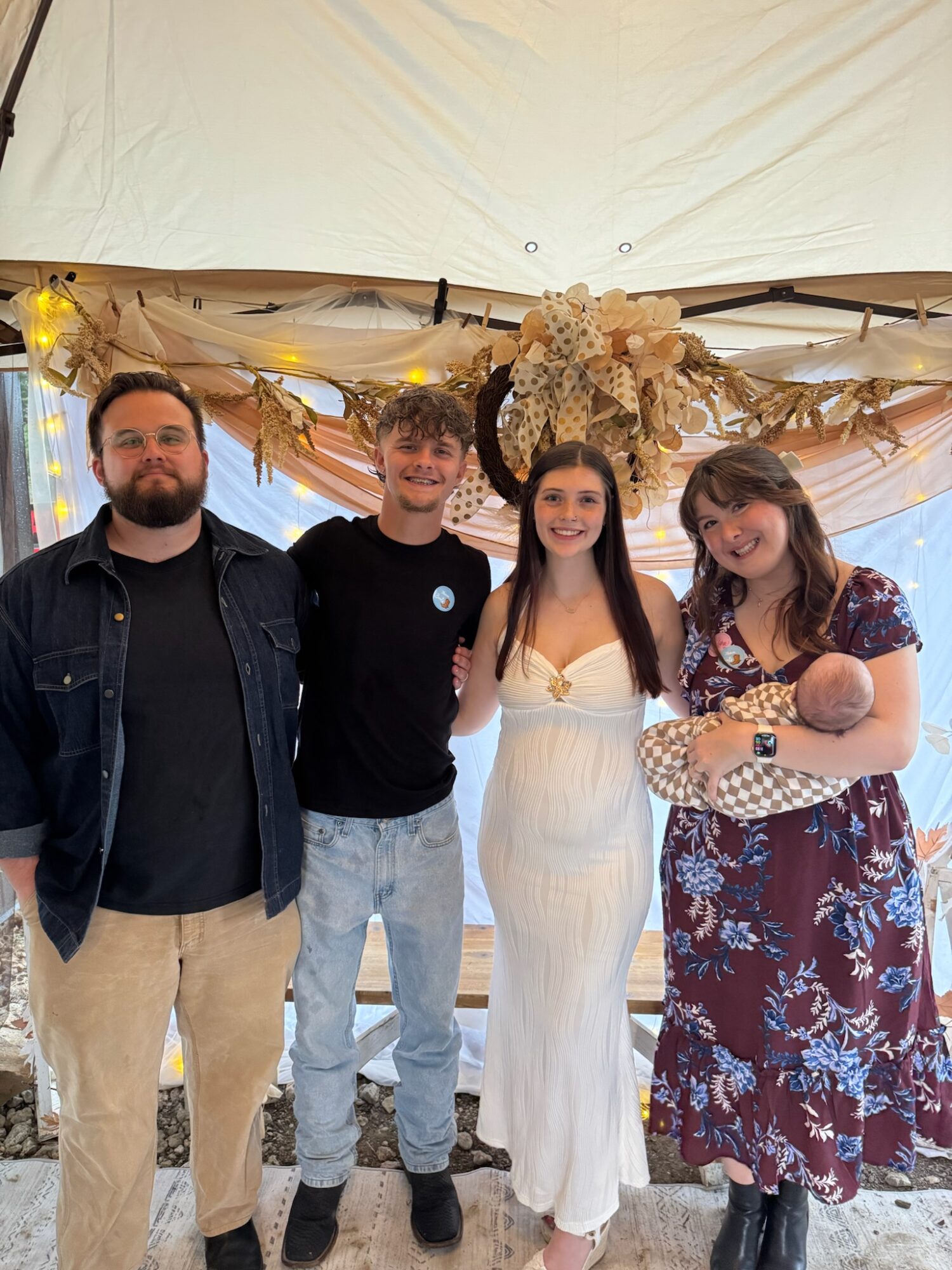 Four people standing together in front of a decorated backdrop with flowers and lights, smiling at the camera.