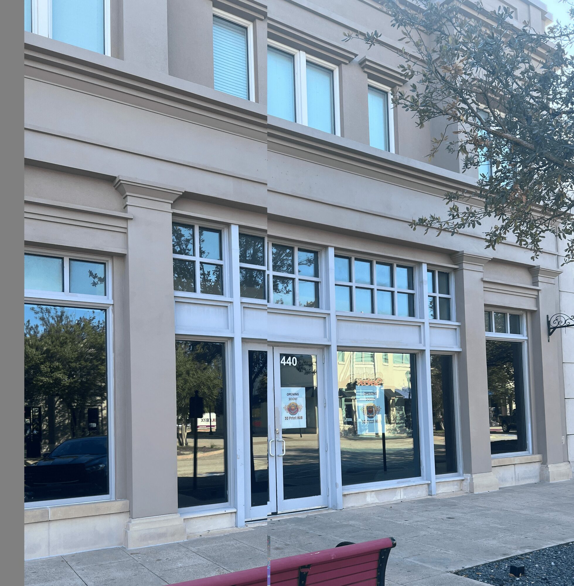 Street-level view of a building with large glass windows and a door, reflecting trees and sky, with a bench in front.