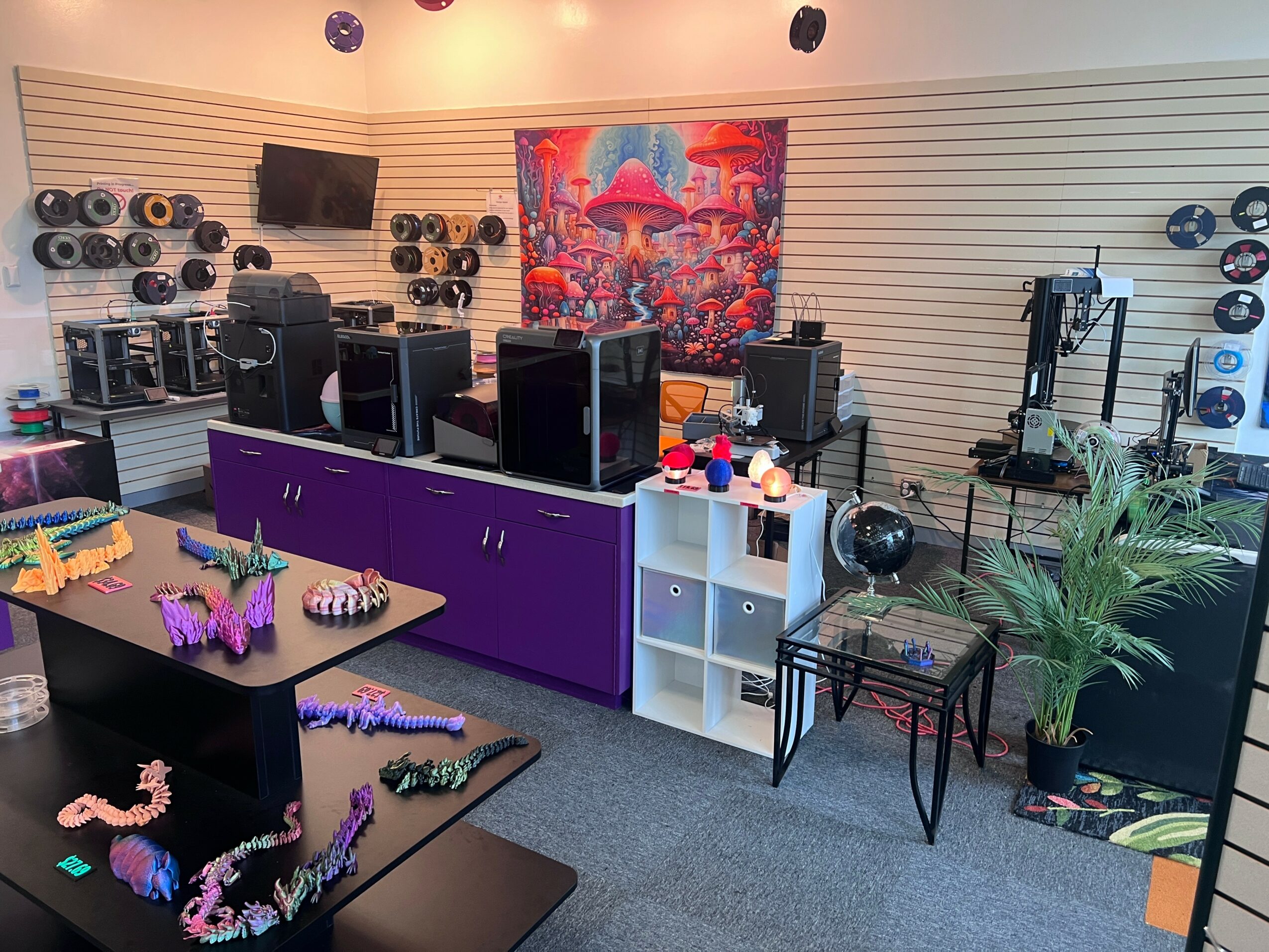 Music studio with computers, speakers, and a colorful wall art, featuring plants and equipment, in a well-lit room.