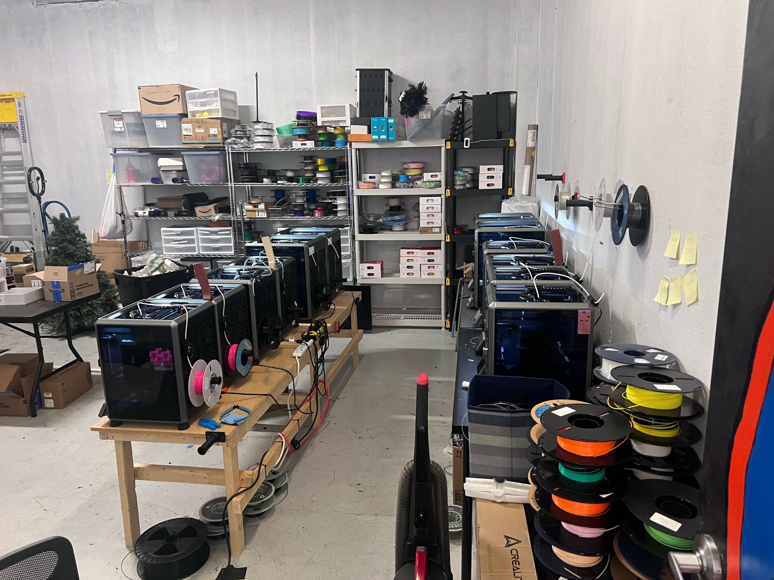 Workshop with 3D printers, shelves with supplies, and tools on the wall, organized workspace, and various equipment.
