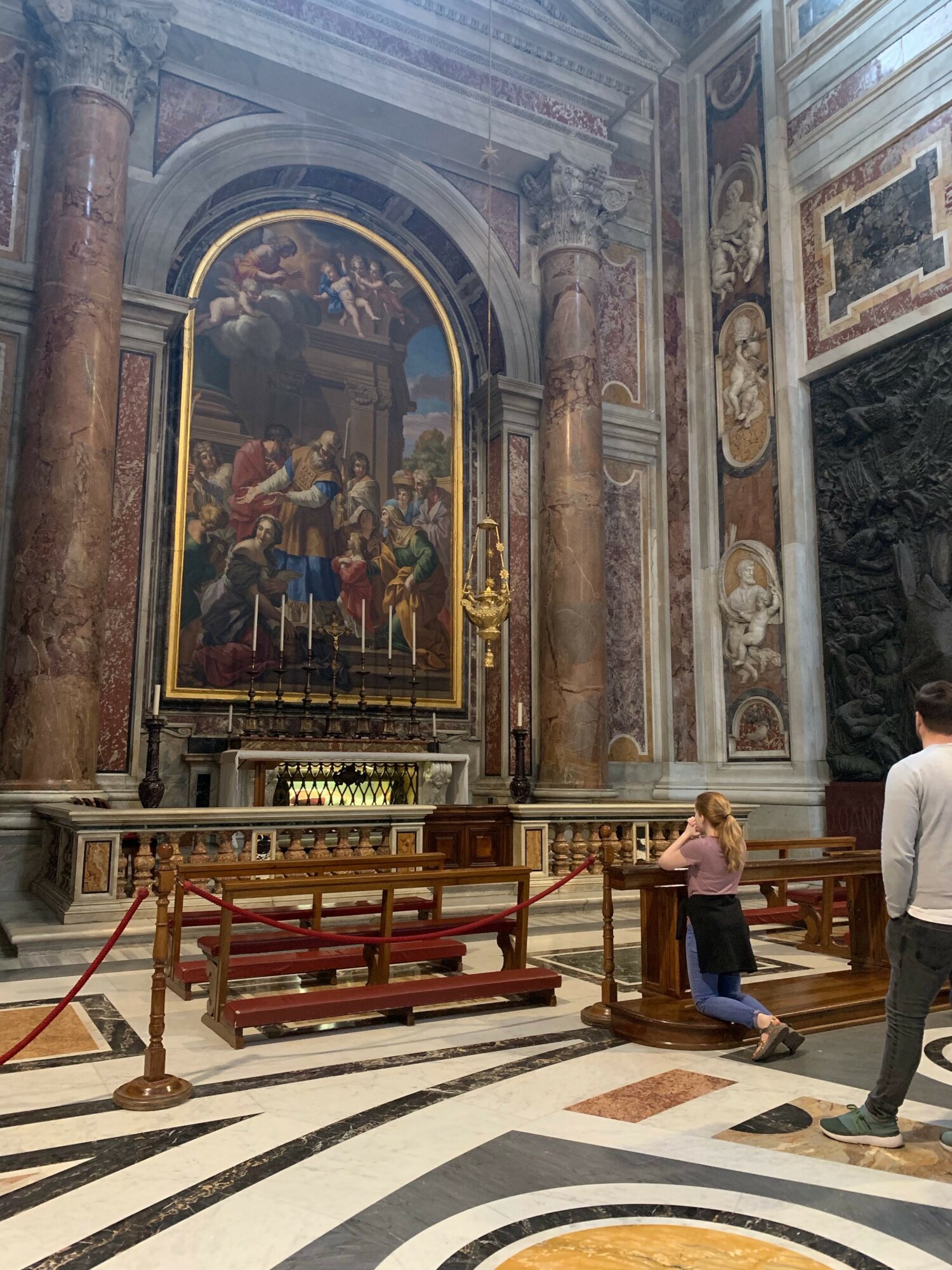 Interior of a church with a large religious painting, ornate columns, and marble flooring. Two people are kneeling and standing near a barrier.