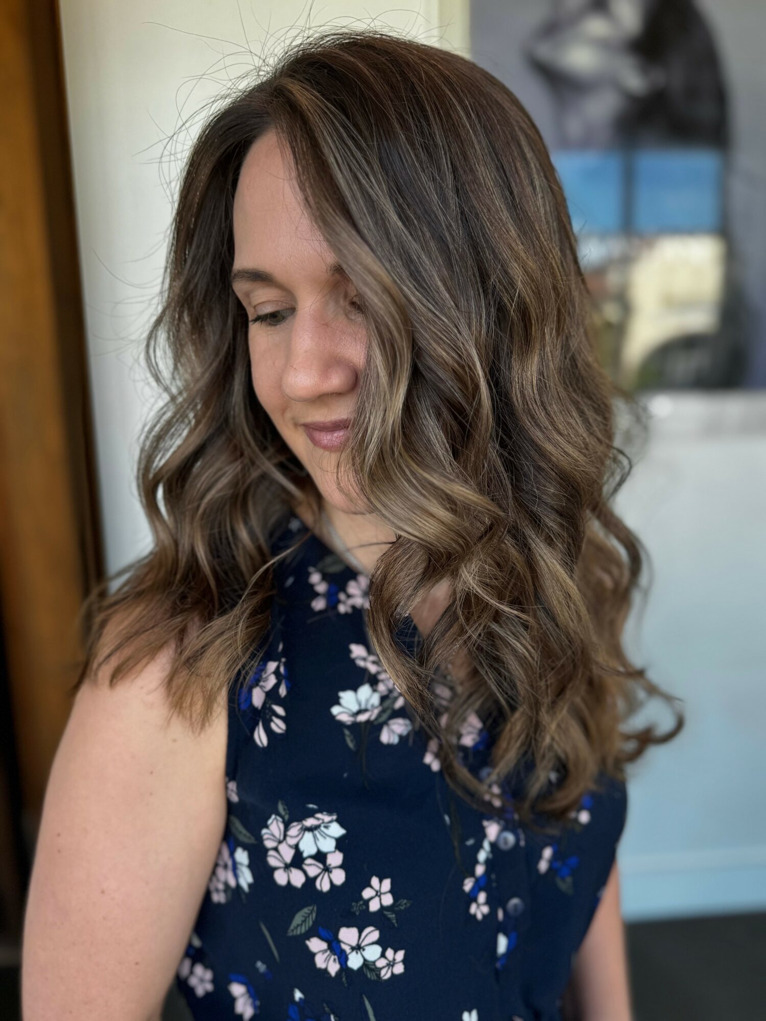 Woman with wavy brown hair looking down, wearing a navy floral dress, indoors with a mirror and window in background.