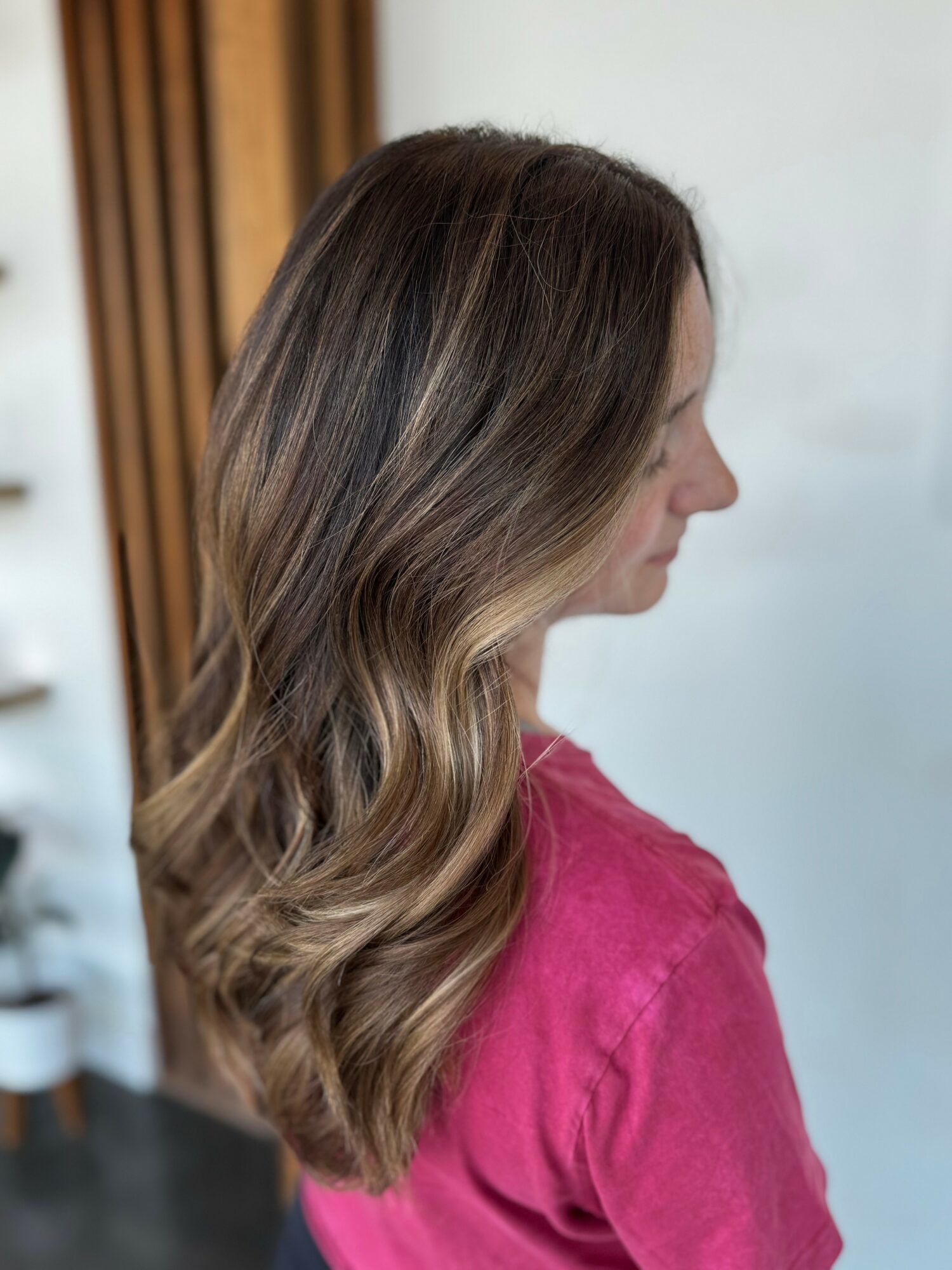Side view of a woman with long, wavy brown hair wearing a pink top, standing indoors against a light background.