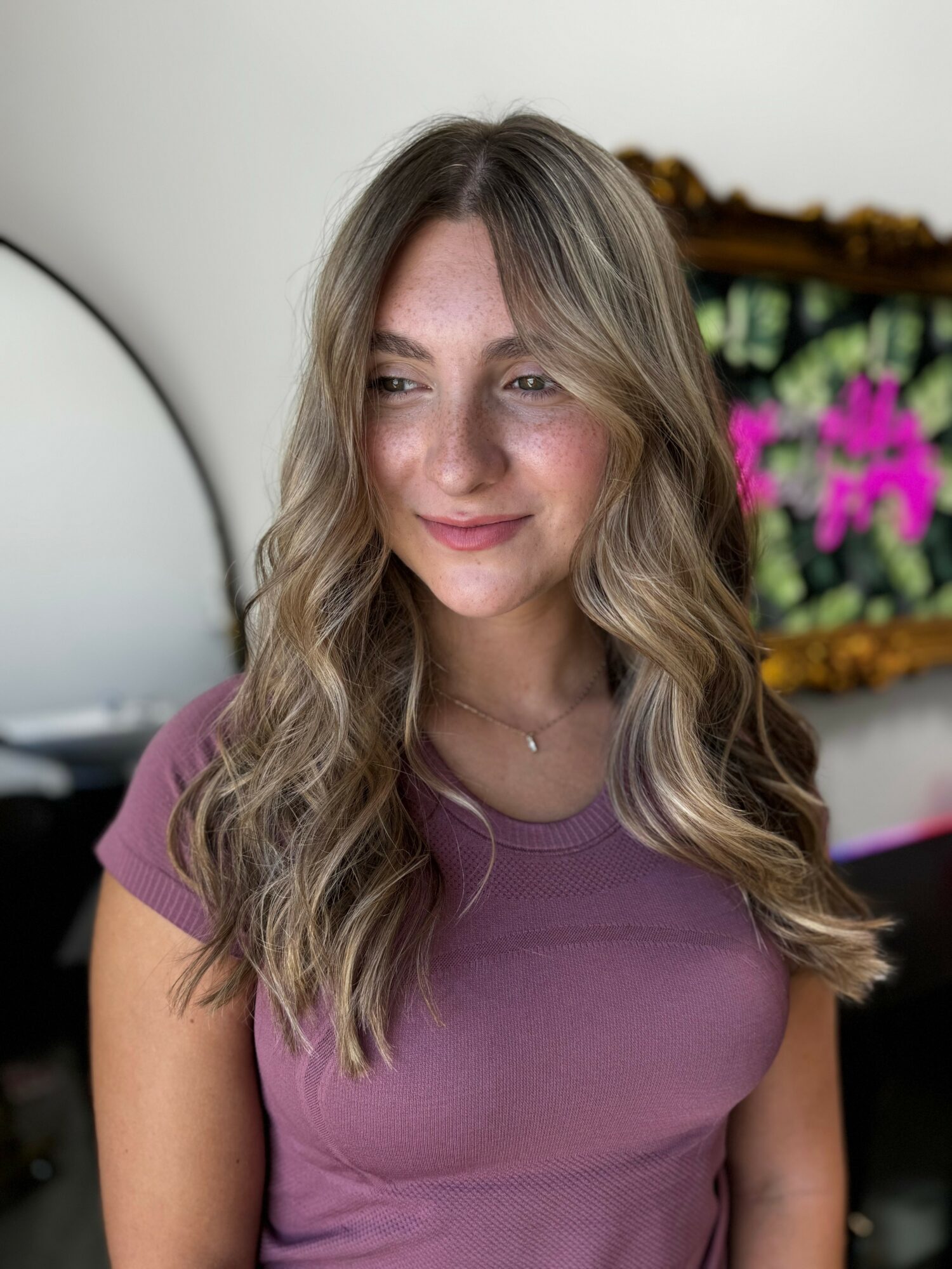 Young woman with long wavy hair wearing a purple shirt, smiling softly, in a room with a mirror and colorful wall art.