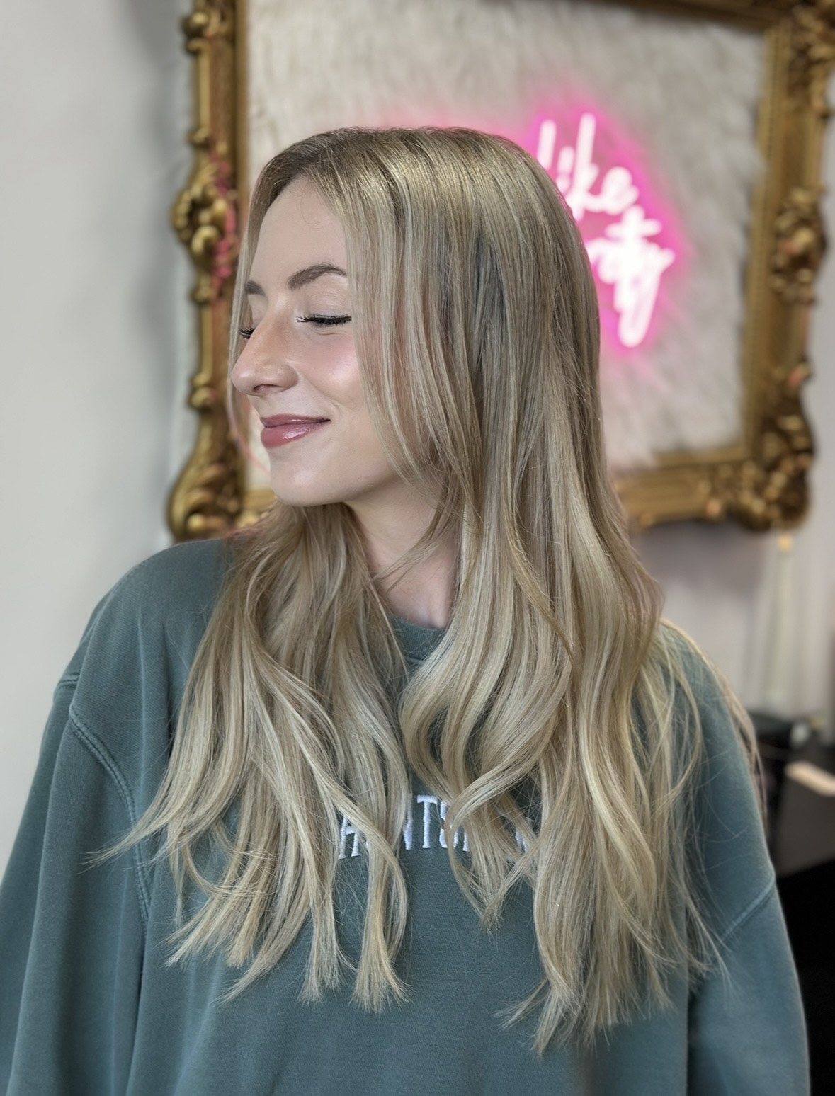 Young woman with long blonde hair smiling with eyes closed, standing indoors with a decorative mirror and pink neon sign behind her.