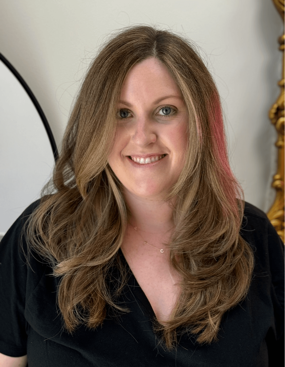 Woman with long, wavy hair smiling, wearing a black top, in a room with a mirror and decorative gold frame