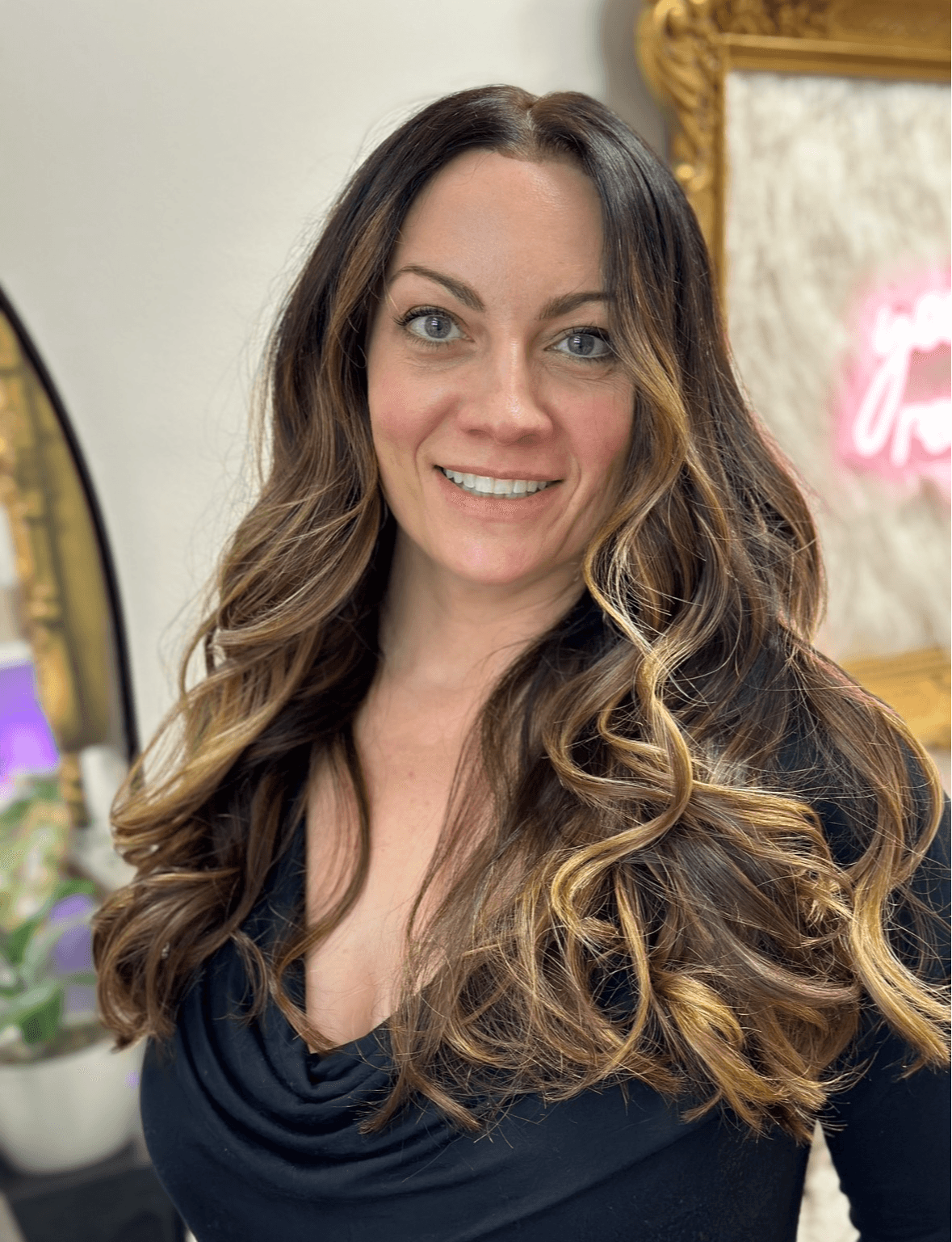 Woman with long, wavy hair smiling, wearing a dark top, in a decorated indoor setting.