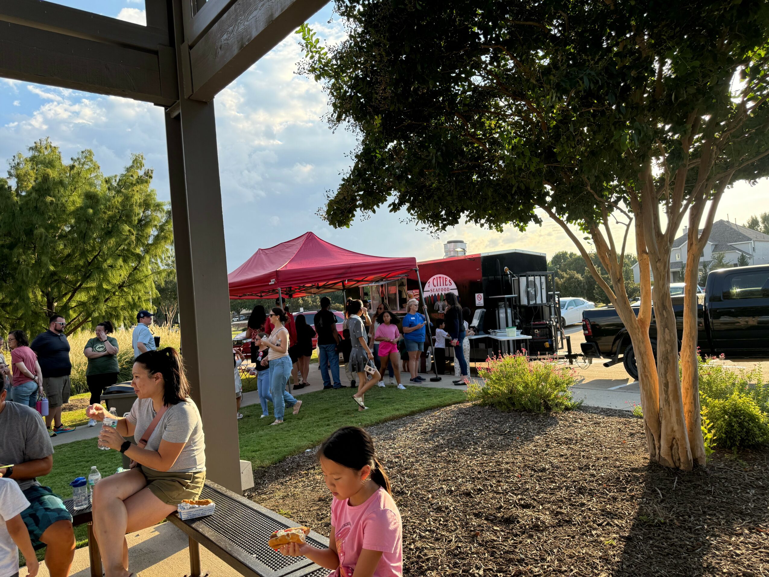 People gathered outdoors near a red canopy, with trees and a parking lot in the background, during daytime.