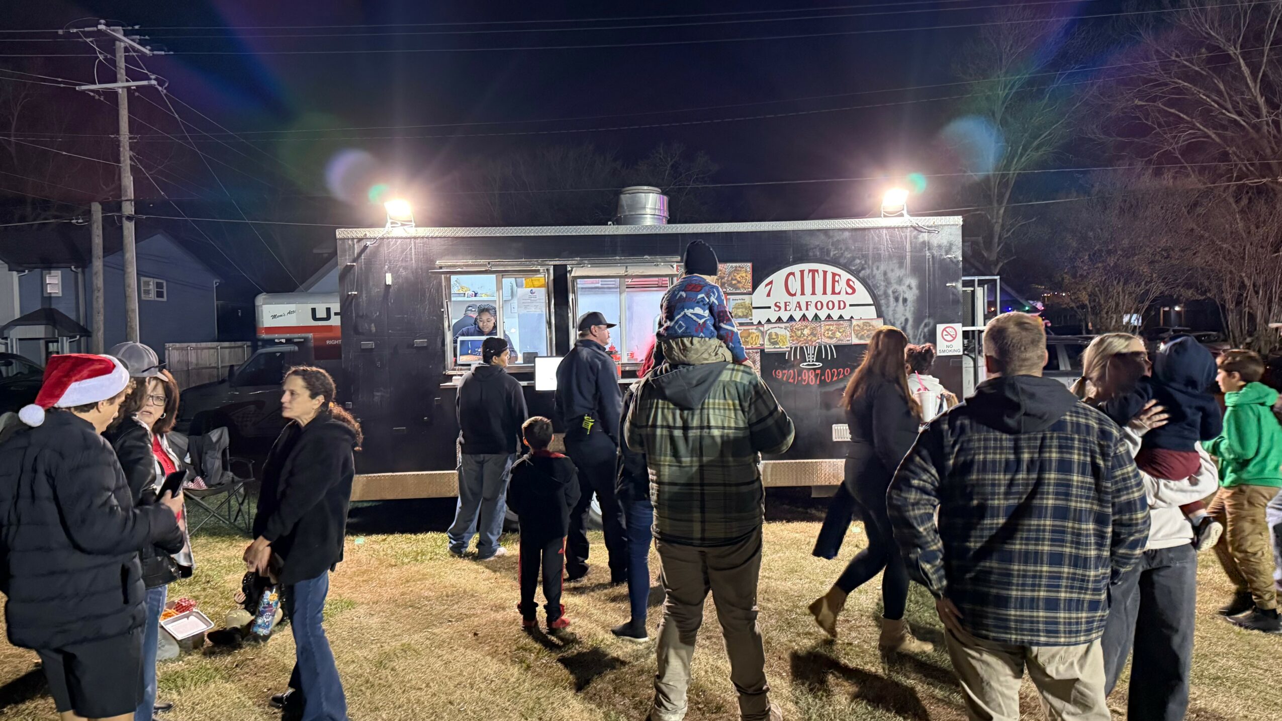 People gather outdoors at night near food trucks with bright lights, some wearing winter clothing and hats.