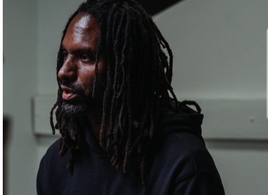 Man with dreadlocks wearing a dark hoodie, looking to the side, indoors.