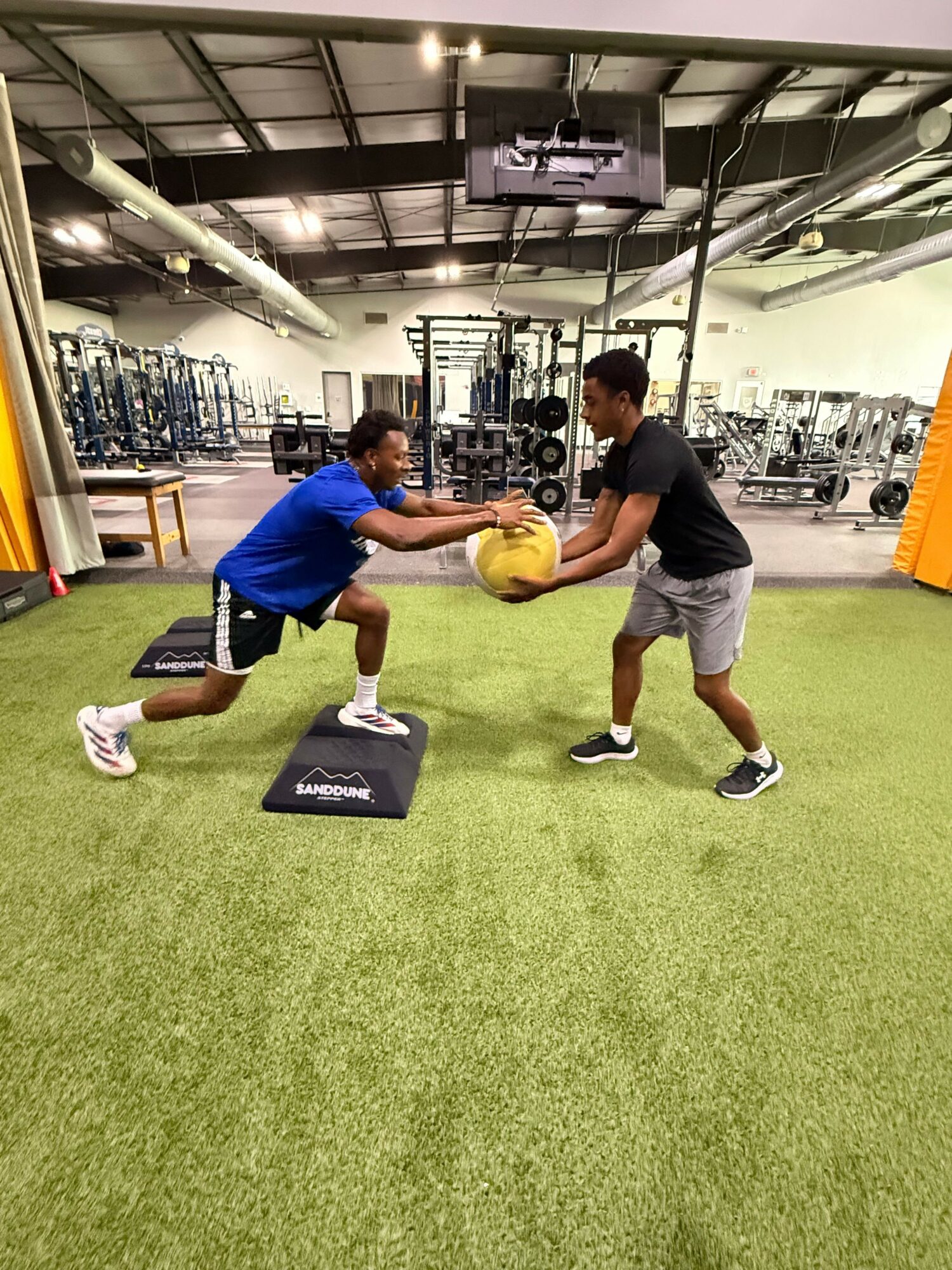 Two people exercising with a yellow ball and a black box in a gym with green turf.