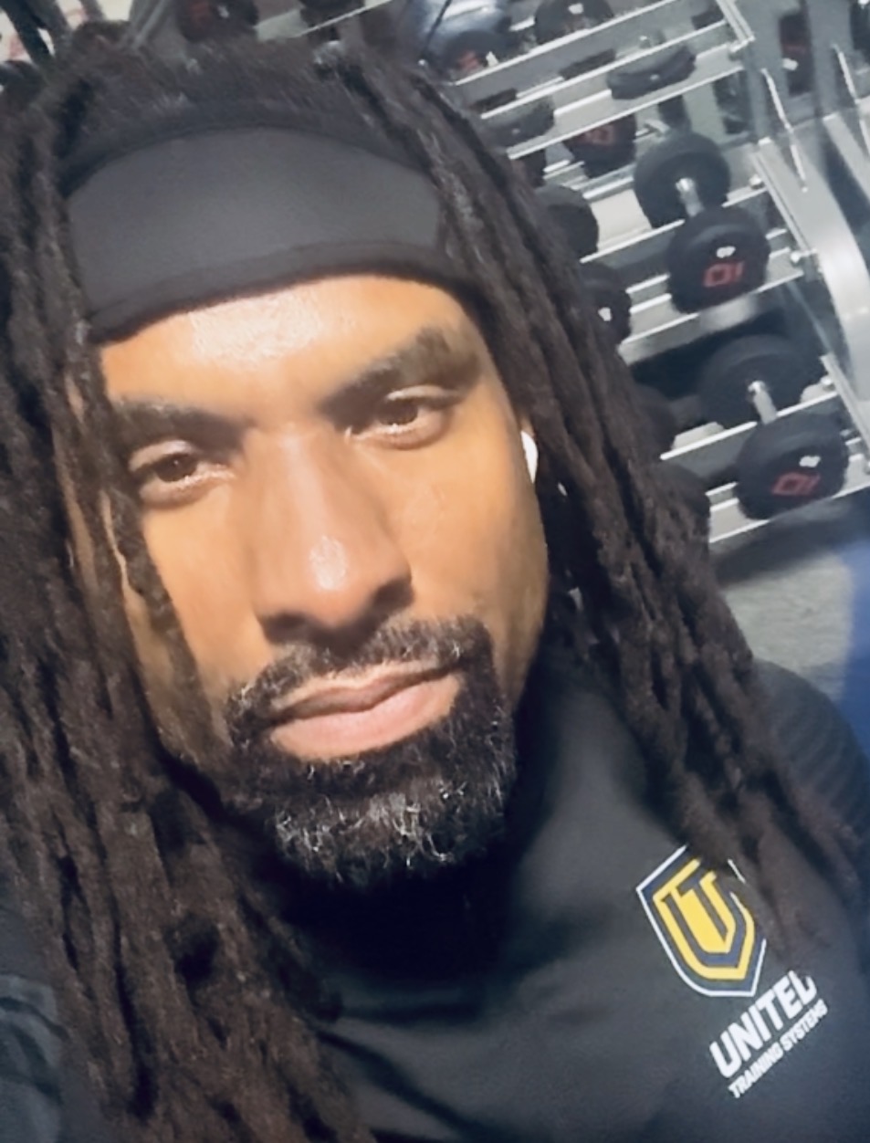 Man with dreadlocks wearing a black headband and sportswear, gym equipment in background.