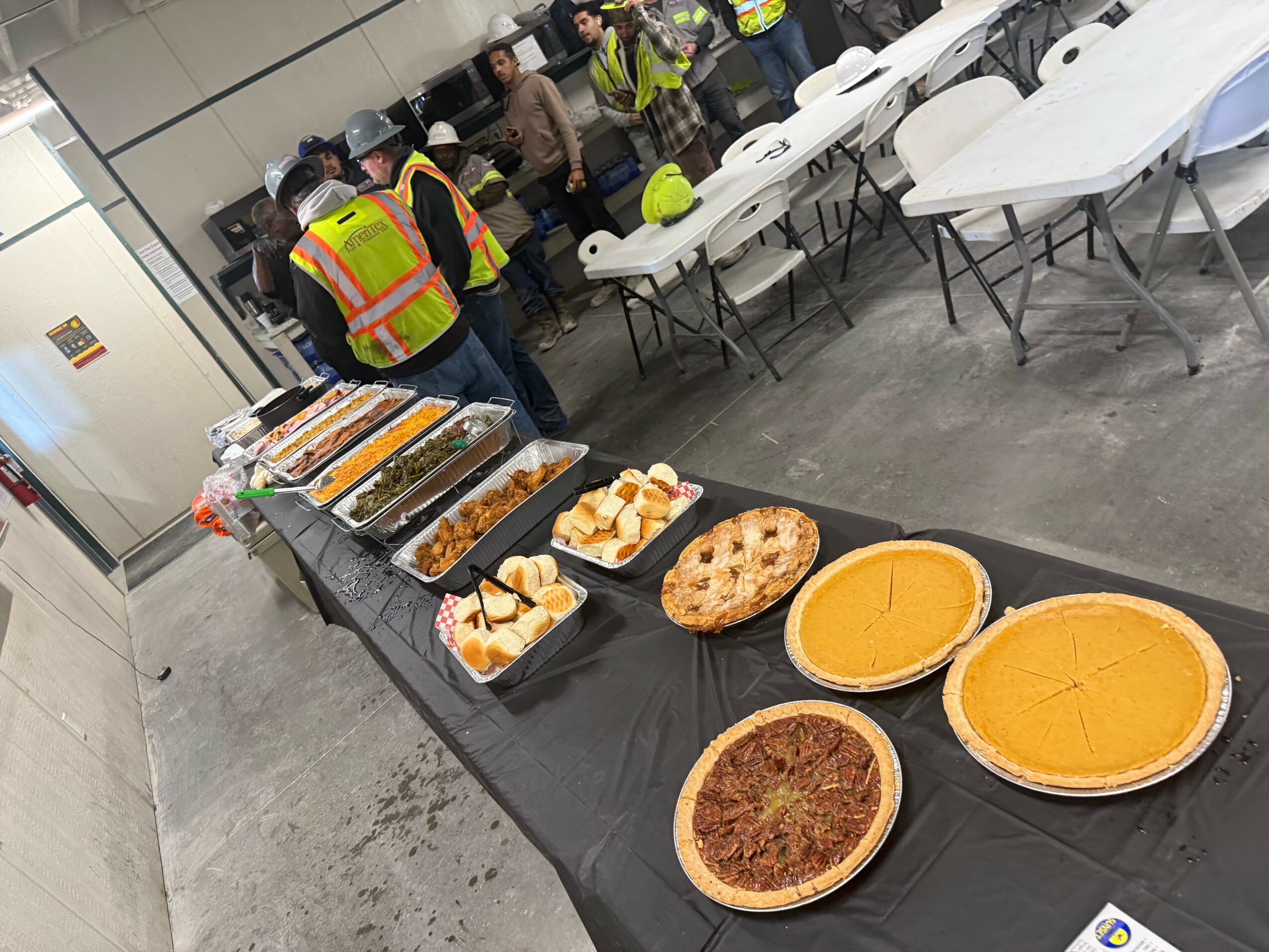 Table with various pies and desserts, with people in safety vests and helmets in the background, and empty chairs nearby.