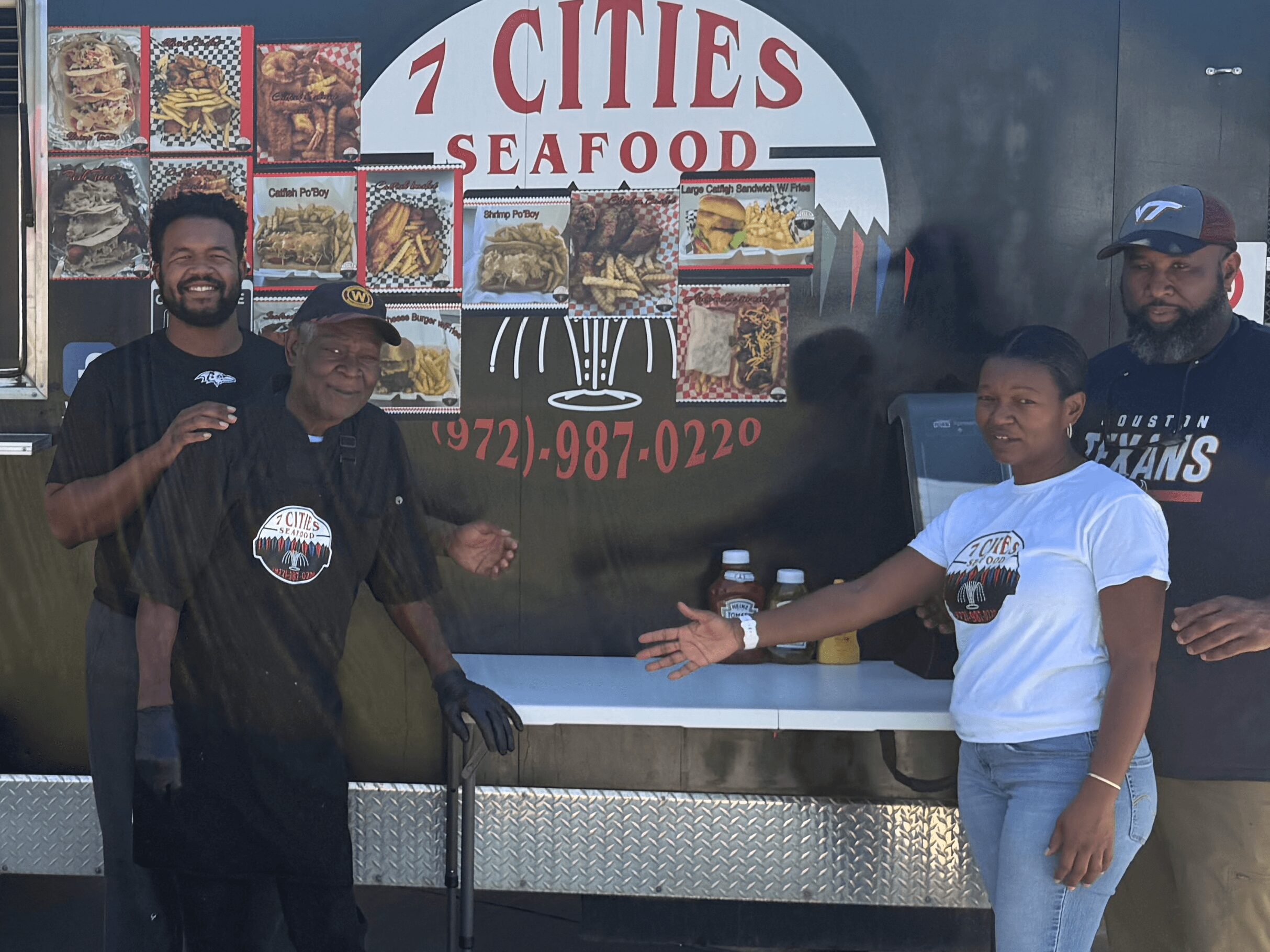 Four people standing in front of a seafood restaurant sign, smiling and gesturing, with food images on the sign.