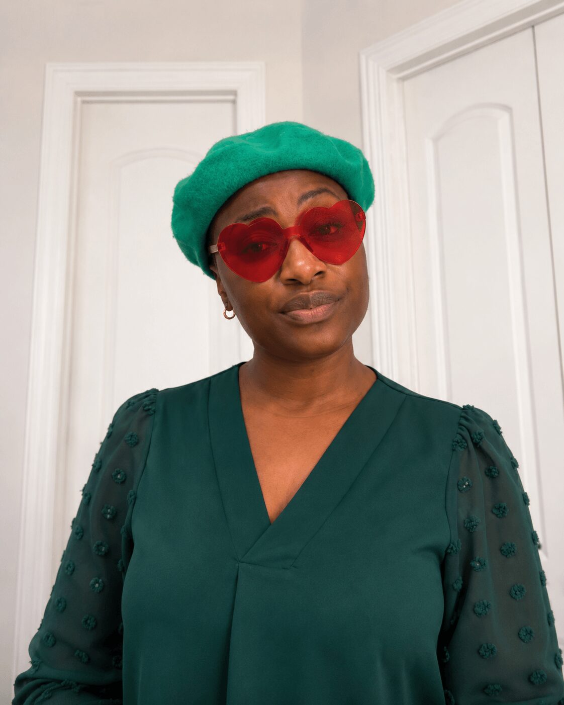 Person wearing a green beret, red heart-shaped sunglasses, and a dark green top with sheer polka-dot sleeves, standing indoors.