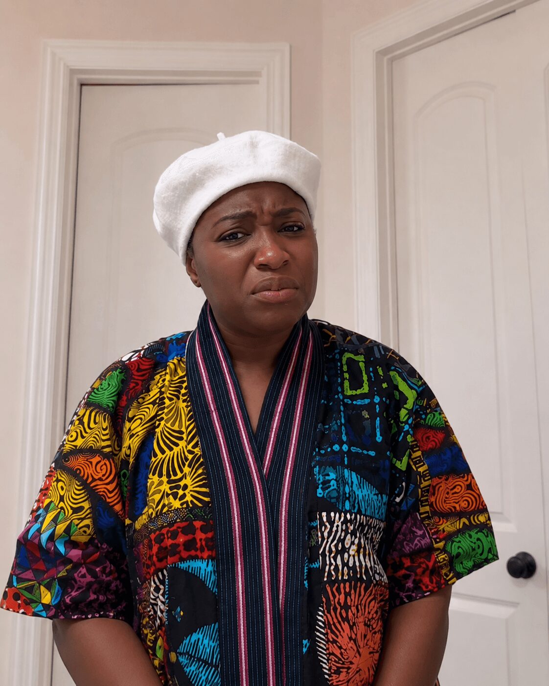 Woman wearing a colorful patterned top and white beret, standing in front of white doors, with a puzzled expression.