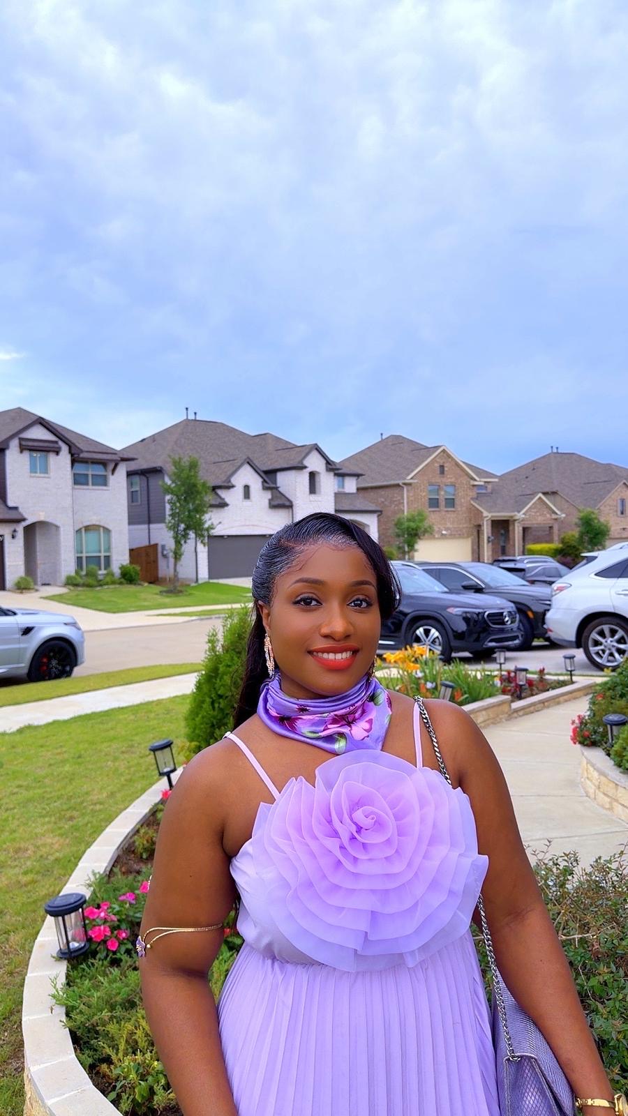 Young woman in a purple dress standing outdoors with houses and cars in the background.