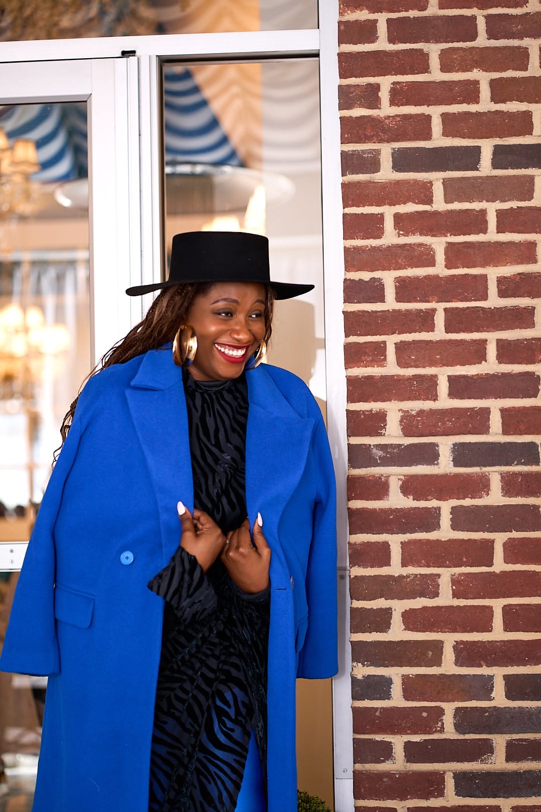 Woman smiling, wearing a black hat and blue coat, standing near a brick wall and glass door.