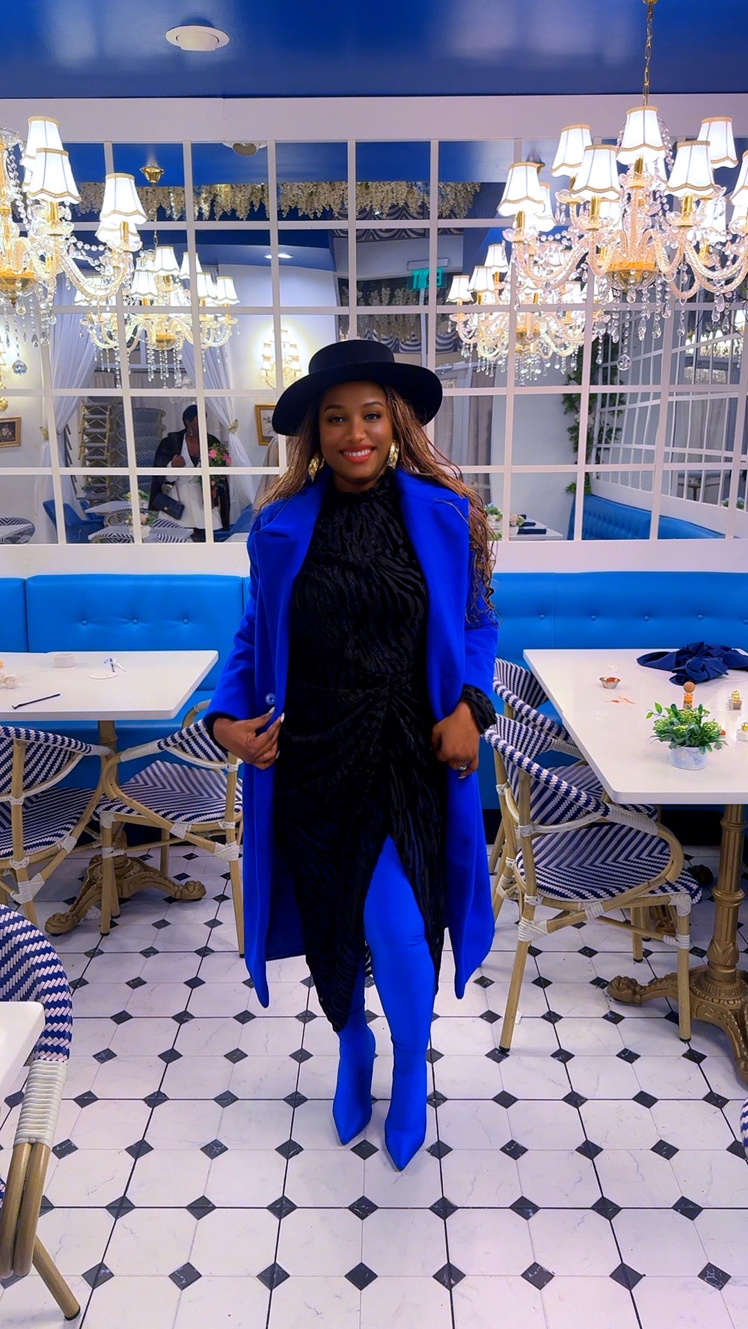 Woman in blue coat and black hat standing in a bright, elegant restaurant with chandeliers and white-tiled floor.
