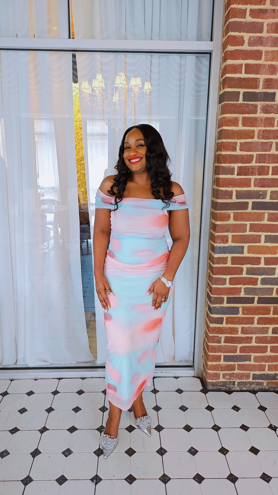 Woman in colorful dress standing indoors near brick wall, smiling, with tiled floor and window behind her.
